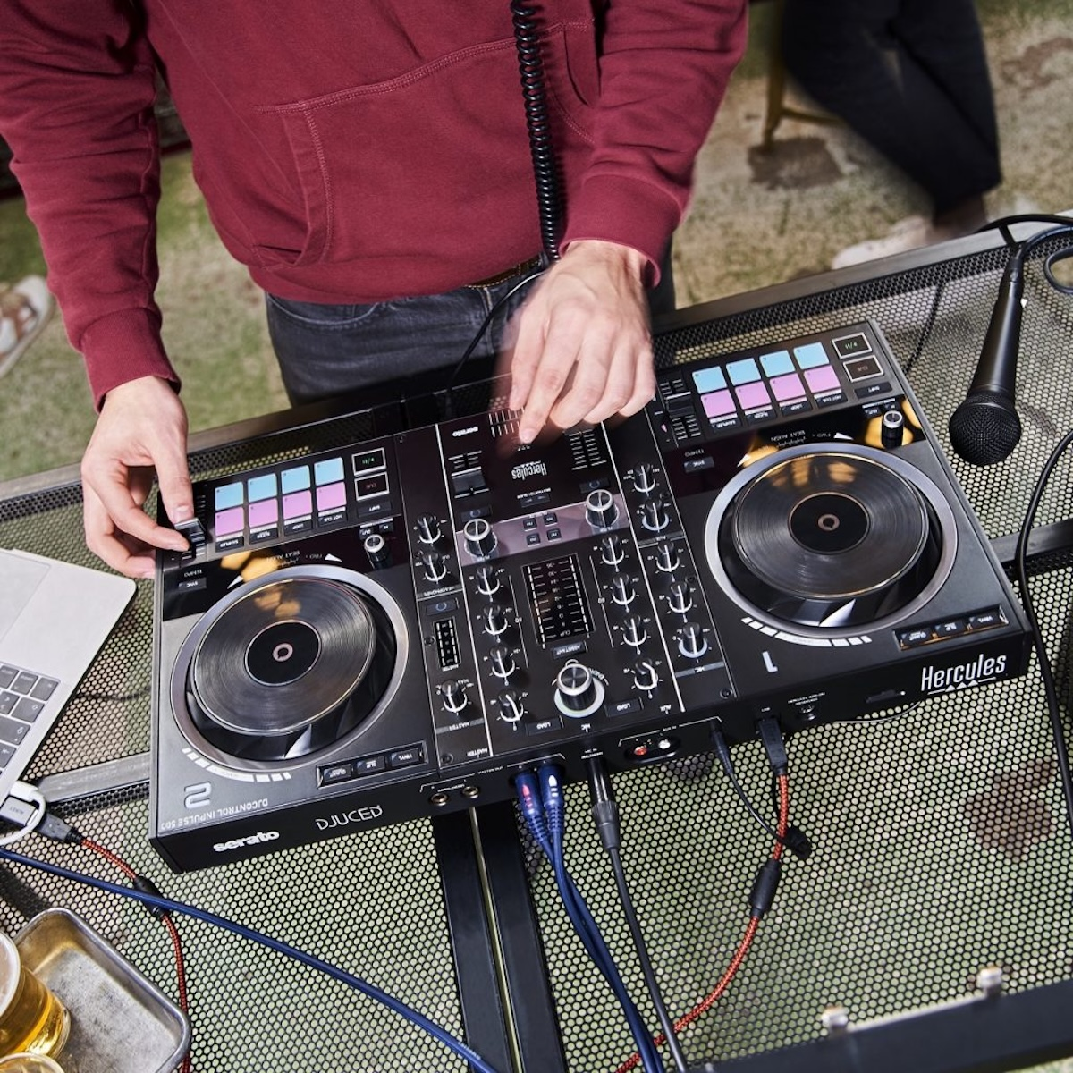 Persoon met een rode jas die een DJ-mixer met microfoon en laptop gebruikt. Drankjes op de voorgrond.