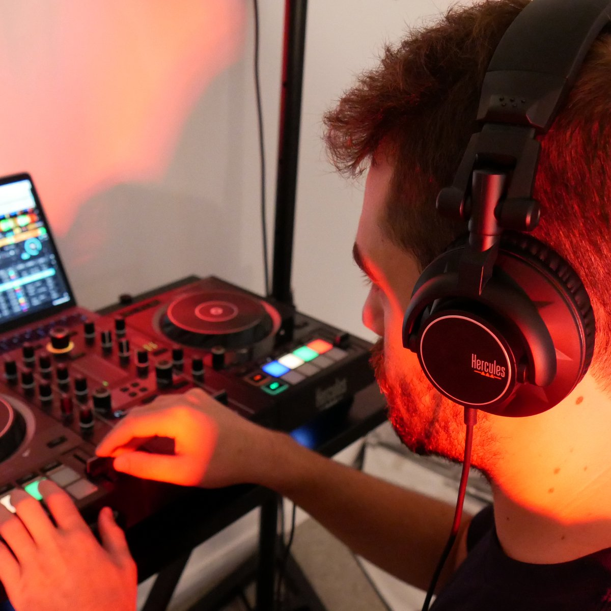 Man met koptelefoon, DJ-setup met laptop, muziek mixen, rode verlichting.