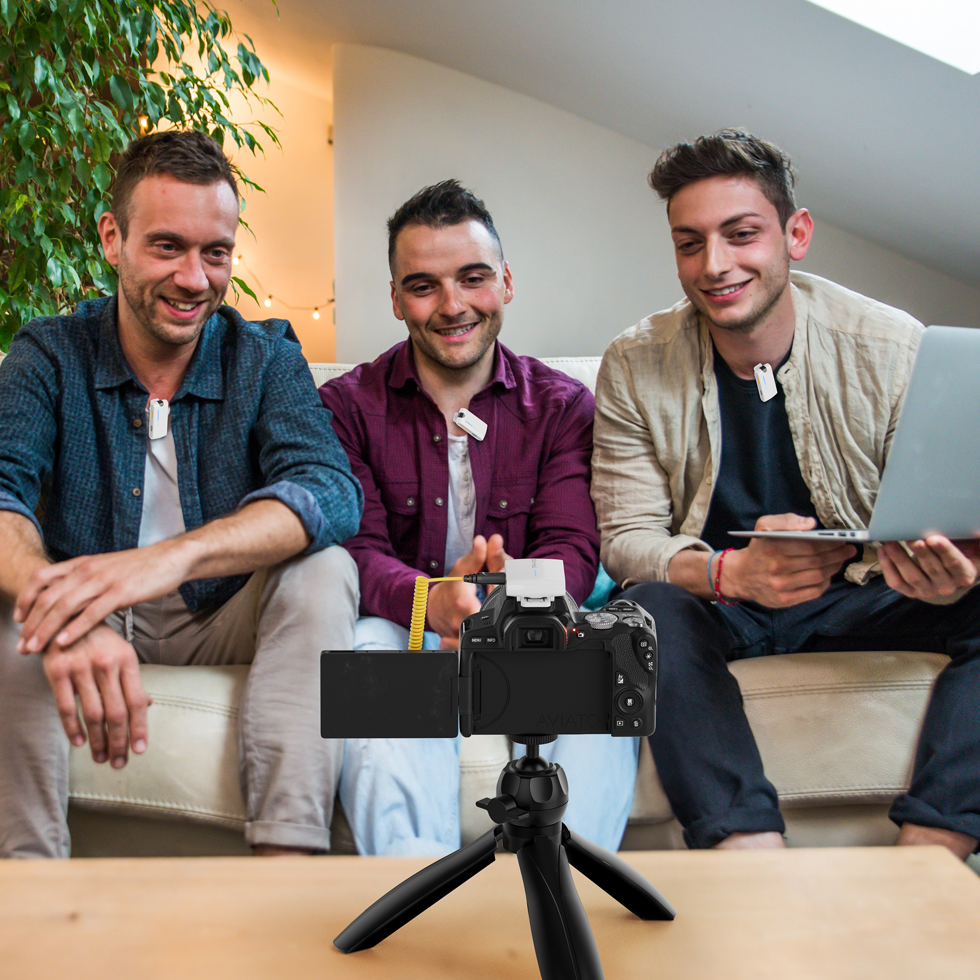 Drie mannen zitten op een bank, elk met een microfoon, voor een camera en een laptop.