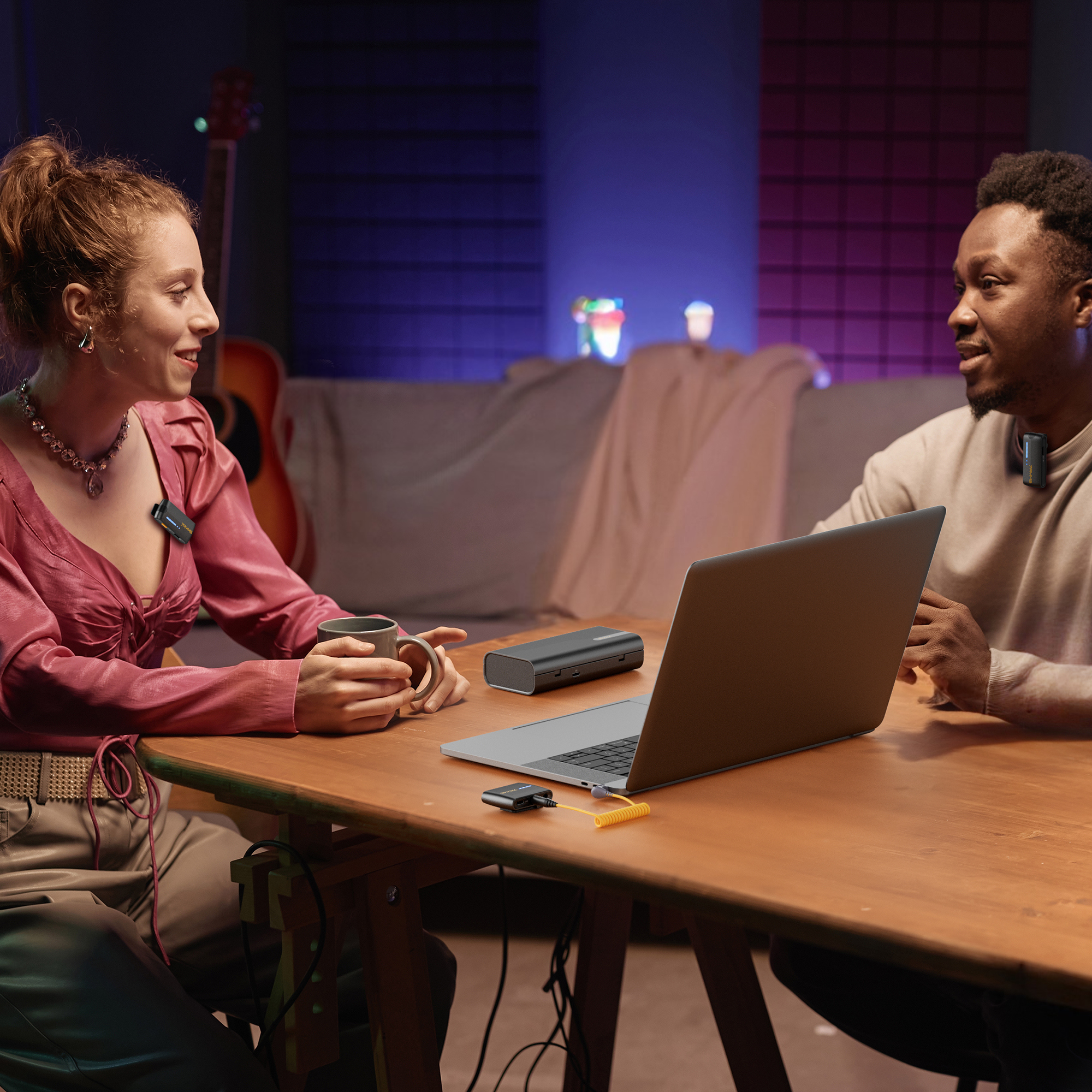 Twee mensen in een kamer met een laptop en microfoons. Een kopje en een oplaadstation zijn ook aanwezig.