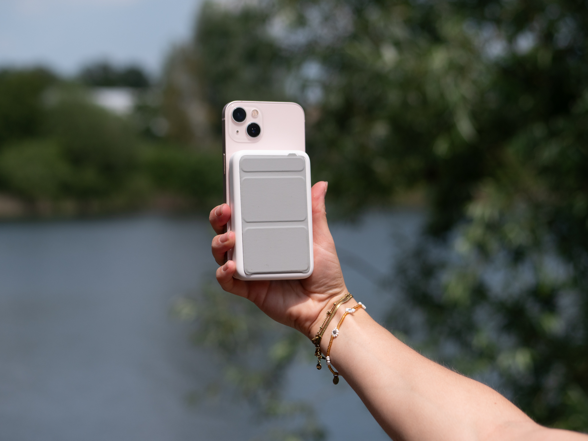 Hand die een telefoon vasthoudt met een wit oplaadaccessoire bevestigd, in de buurt van water en bomen.