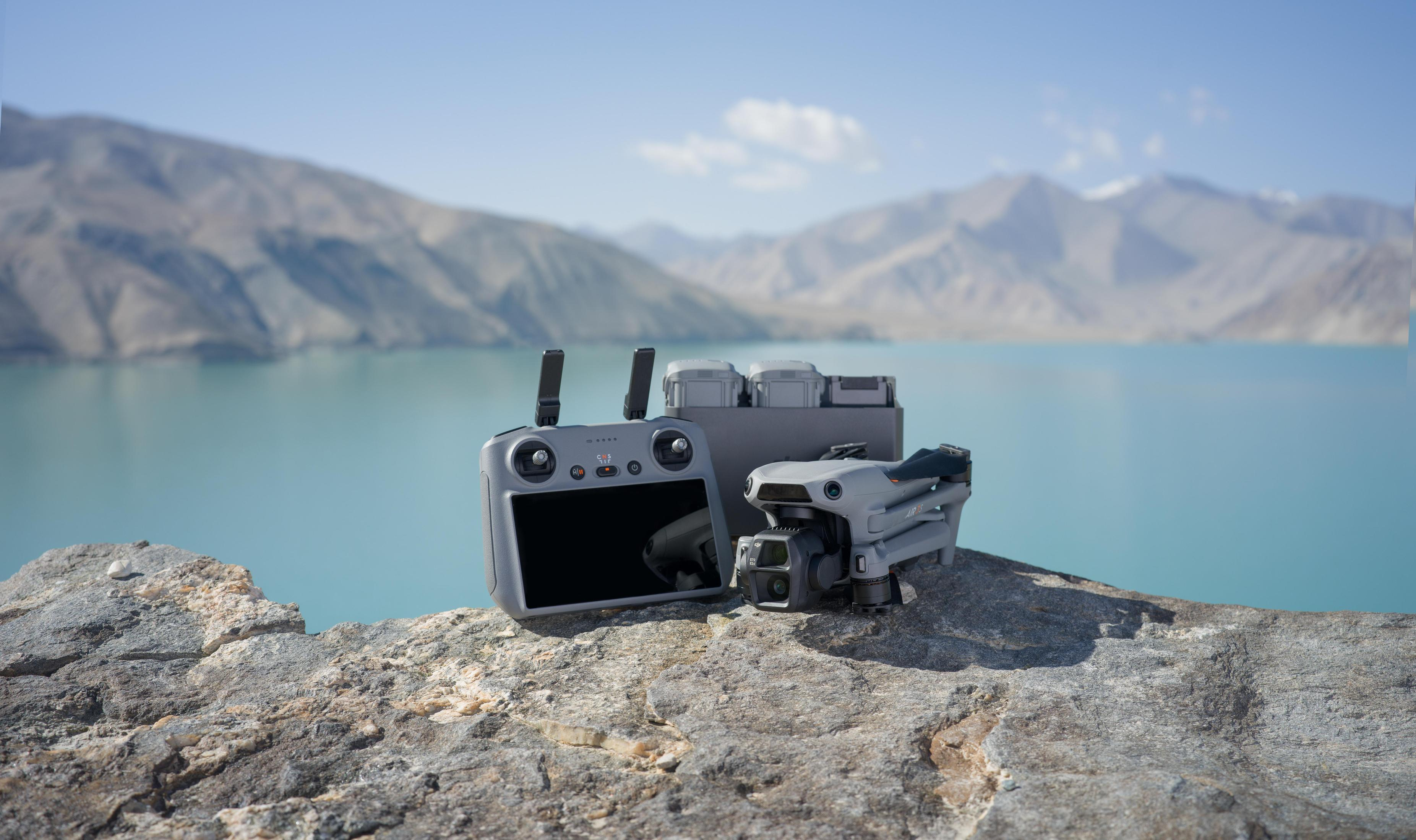 Dron, kontroler i baterie na skale, z jeziorem i górami w tle.