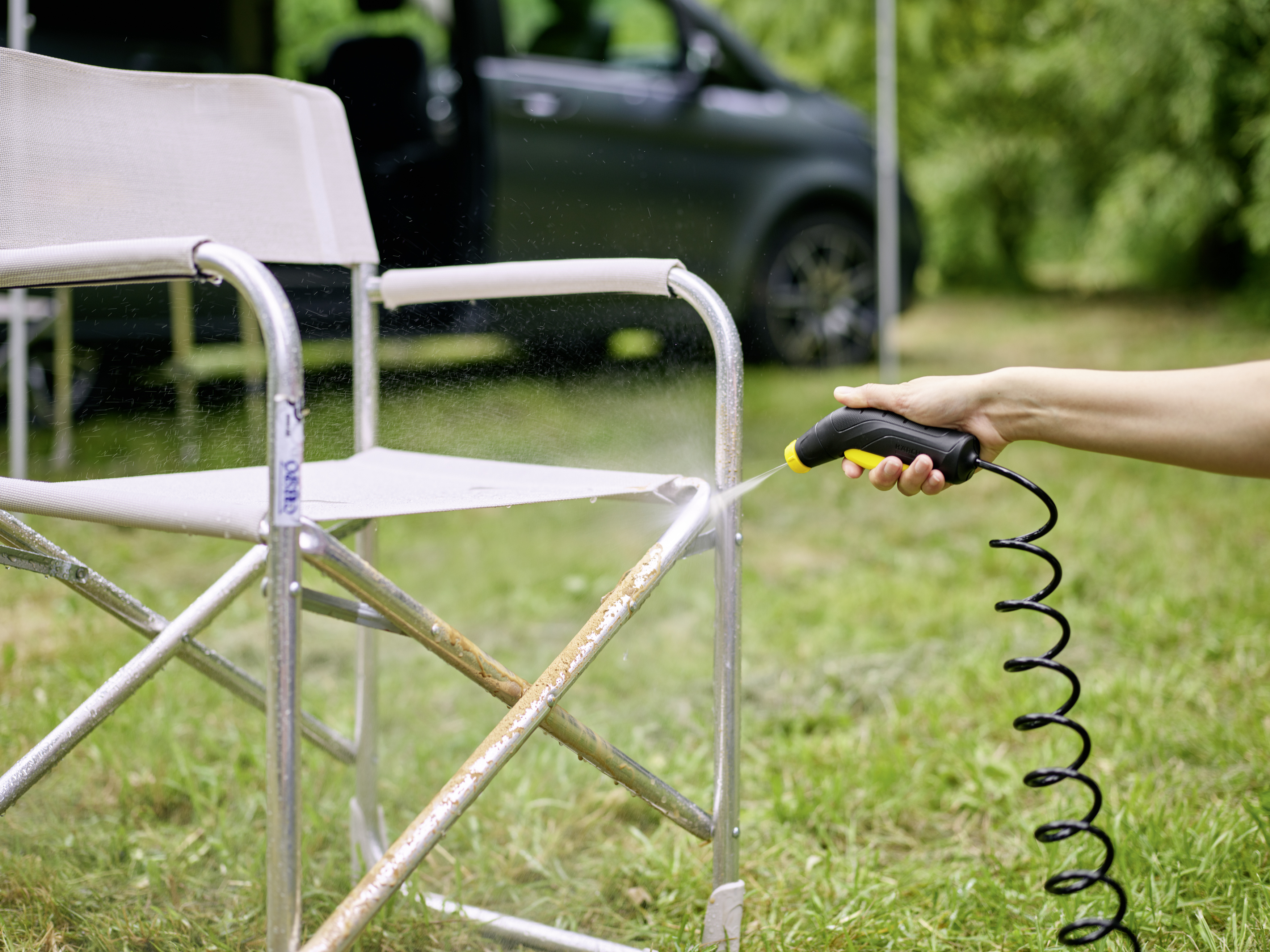 Personne pulvérisant de l'eau sur une chaise pliante. Herbe verte et véhicule en arrière-plan.