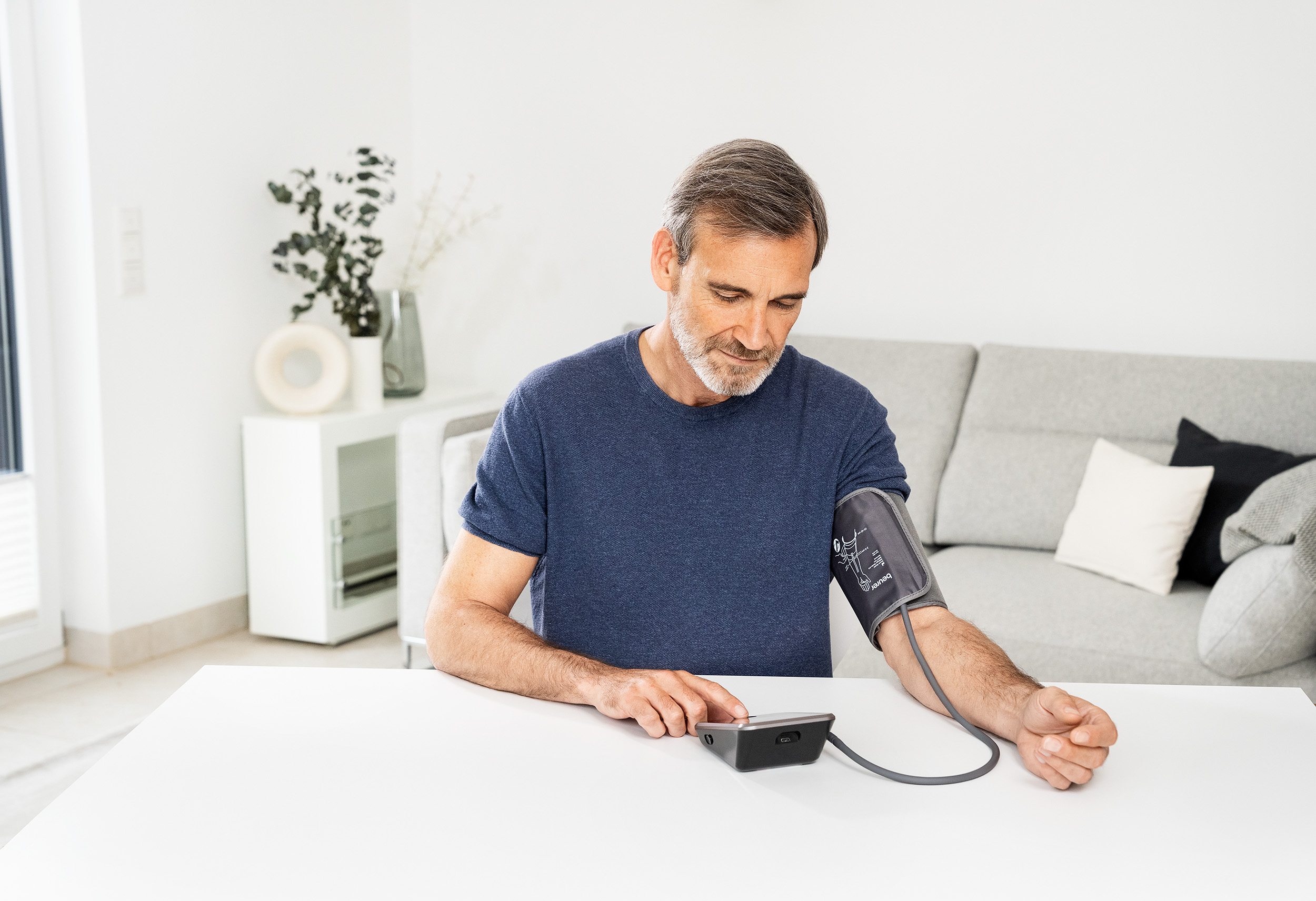 A man checking his blood pressure at a table. He is wearing a blue shirt. There is a sofa in the background.