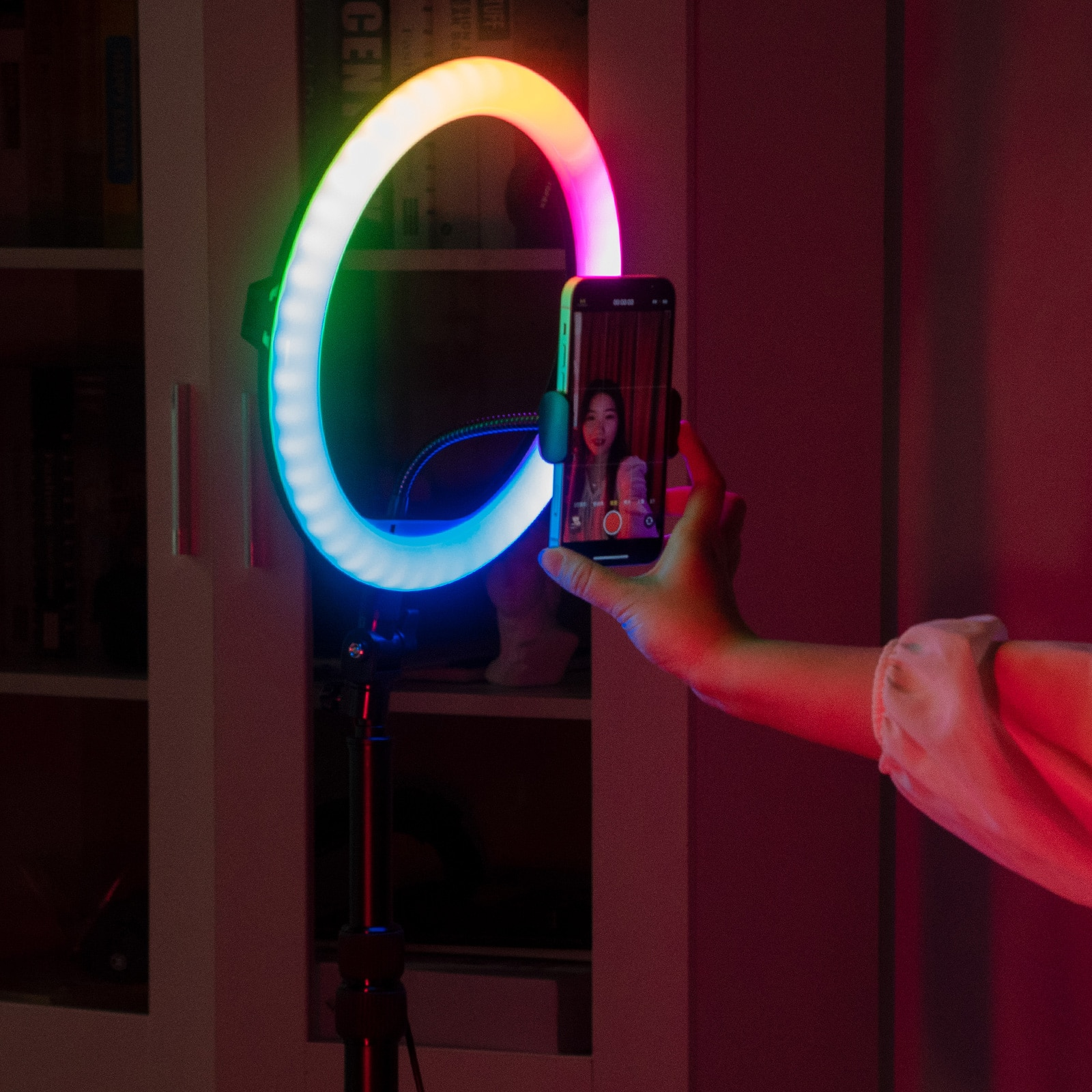 A hand holds a phone in front of a ring light. The light is multi-colored, illuminating the face on the phone.