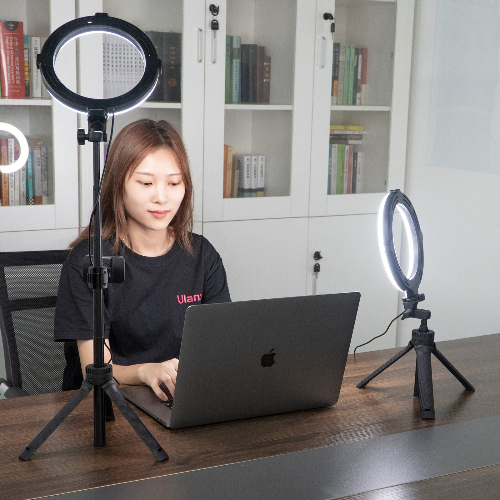Een vrouw die een laptop gebruikt met ringlampen op statieven. Boekenkasten op de achtergrond.