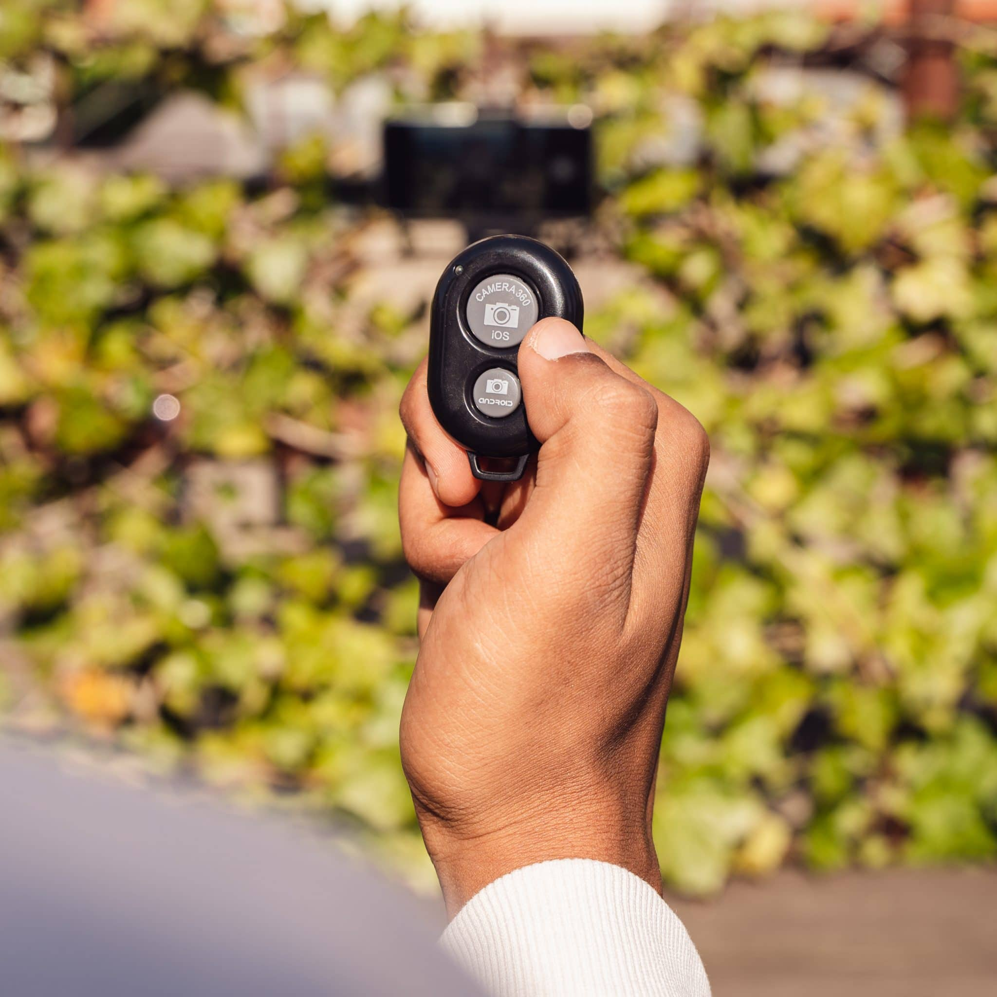 Hand holding a black remote with camera and iOS buttons, leaves in the background.