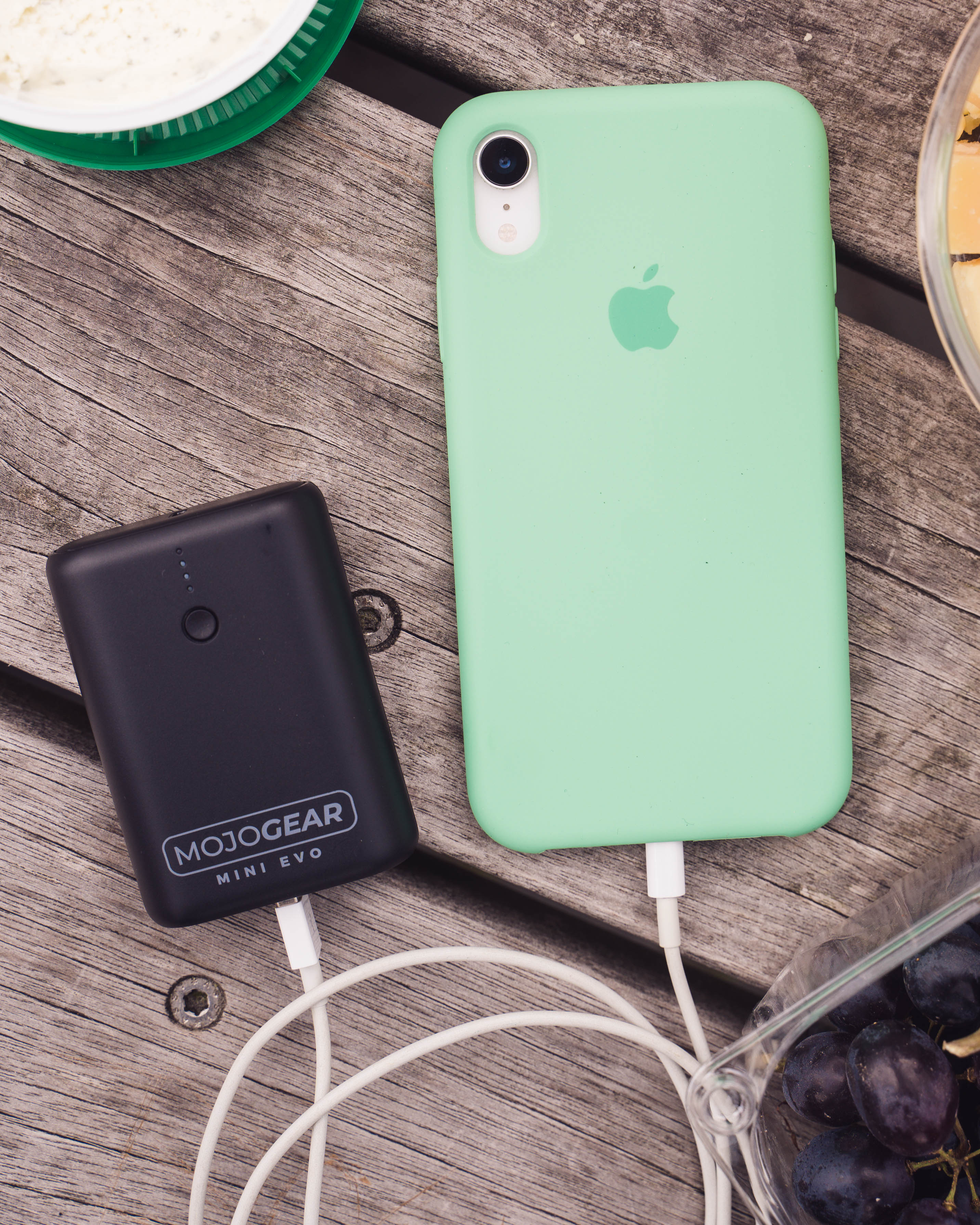 A mint green phone charging a black MojoGear power bank, on a wooden table, with grapes.