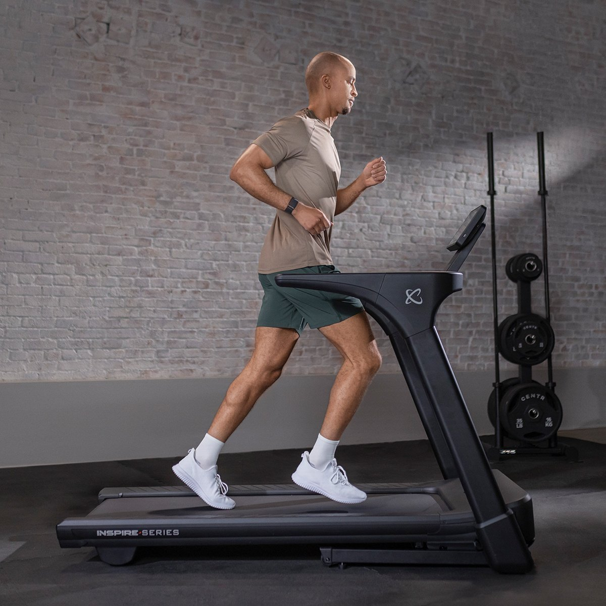 A man runs on a treadmill in a gym. He wears athletic clothes. Background: a brick wall and weight rack.