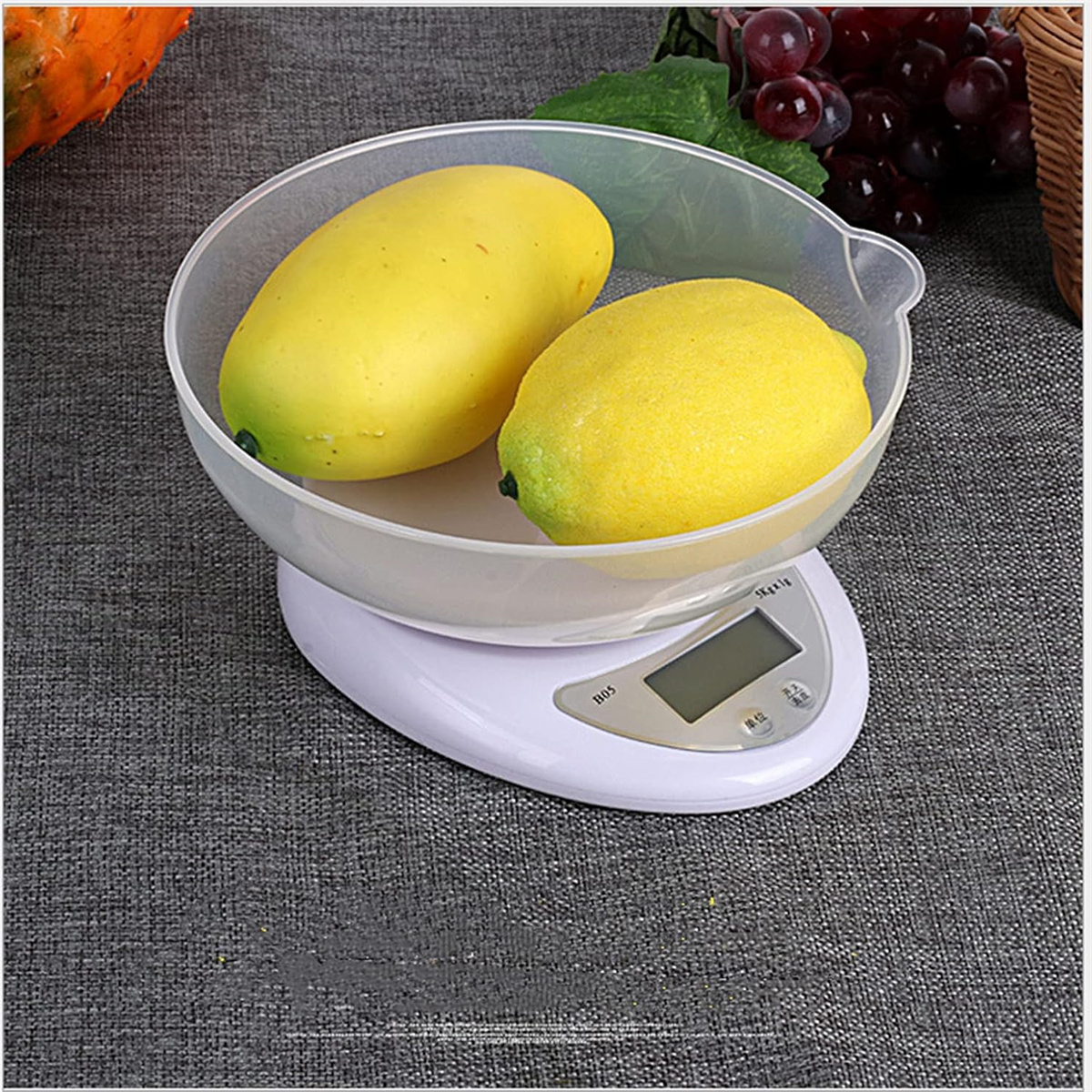 Zwei gelbe Mangos in einer durchsichtigen Schüssel auf einer weißen Waage. Grauer Hintergrund.