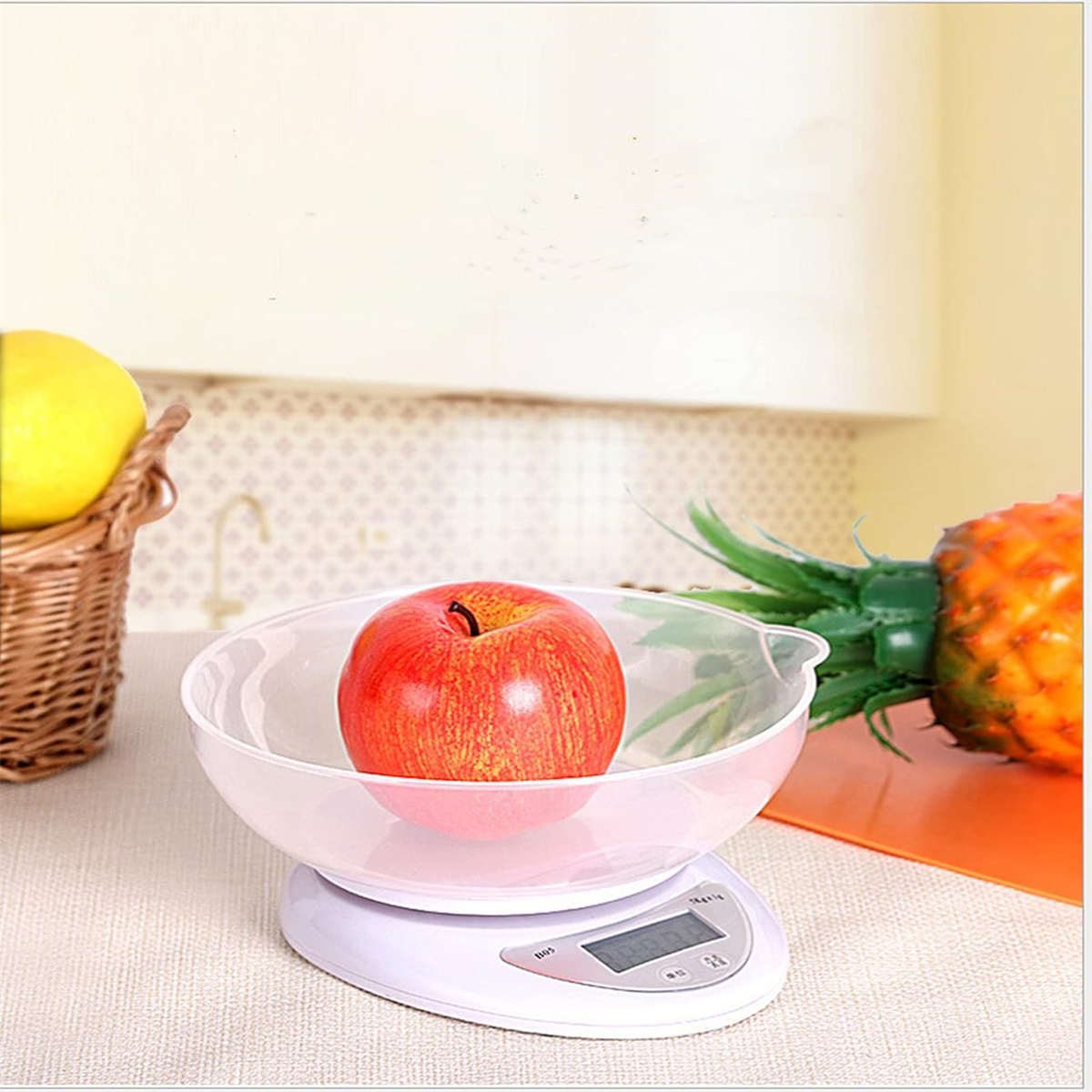 Ein Apfel in einer durchsichtigen Schüssel auf einer Waage. Im Hintergrund ein Korb mit Äpfeln und eine Ananas.