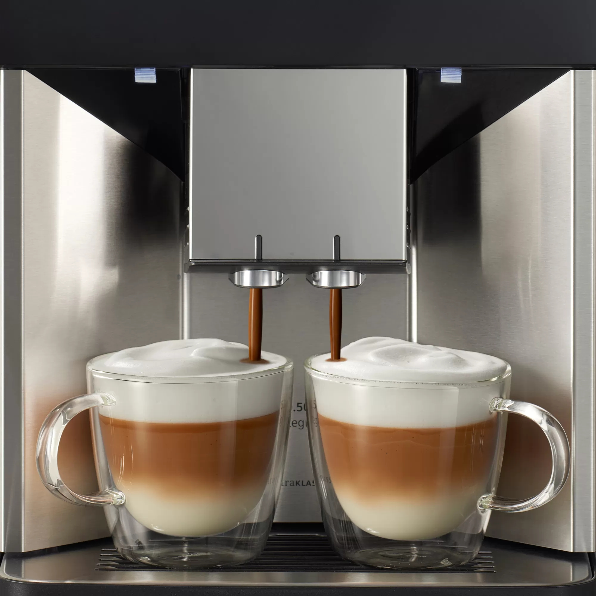 Kaffee wird aus einer modernen Kaffeemaschine in zwei Glasbecher gegossen. Der Kaffee hat Schichten aus weißem Schaum und Farbe.