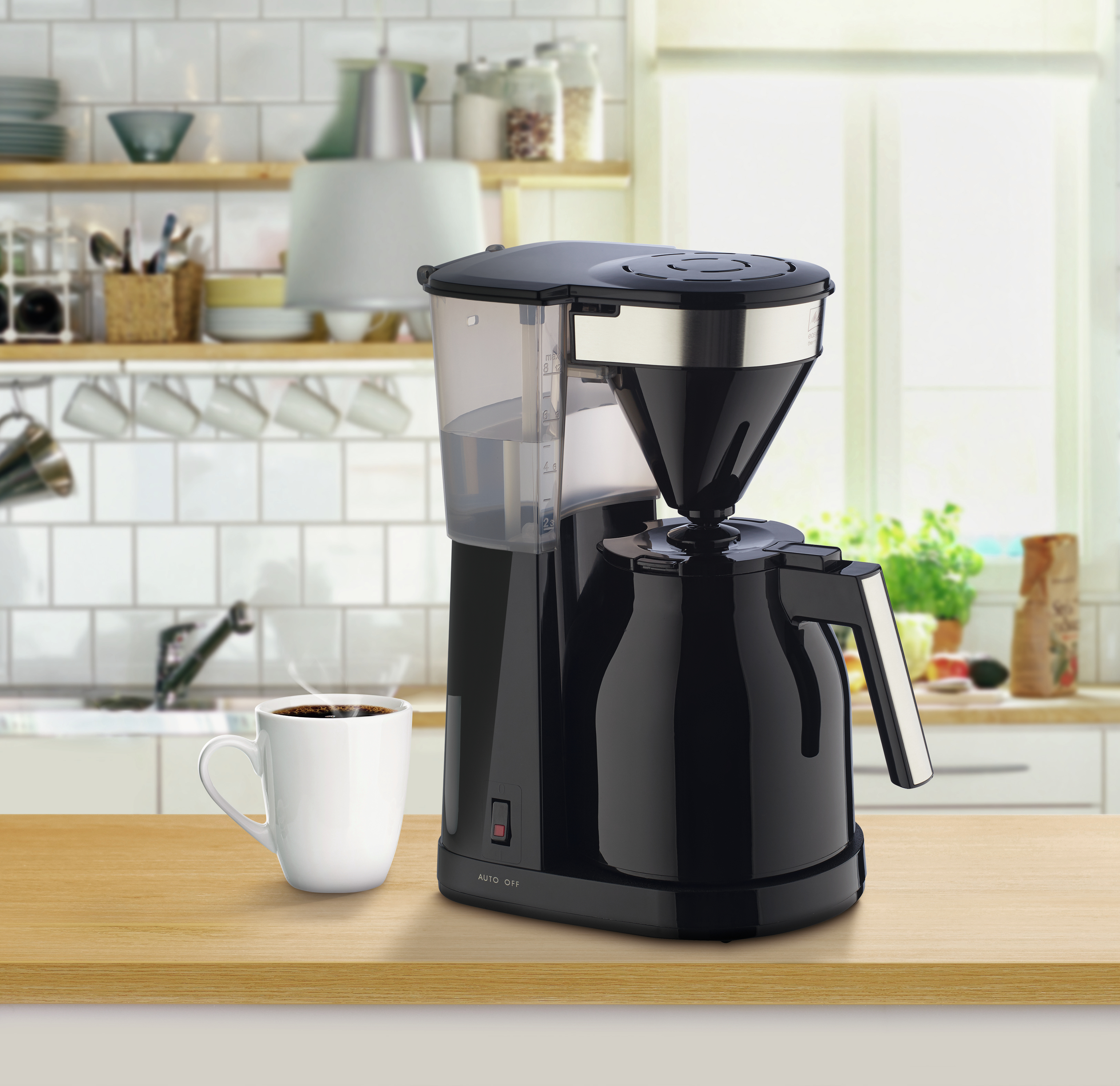 Czarny ekspres do kawy z białą filiżanką kawy na drewnianym stole. Tło to kuchnia.