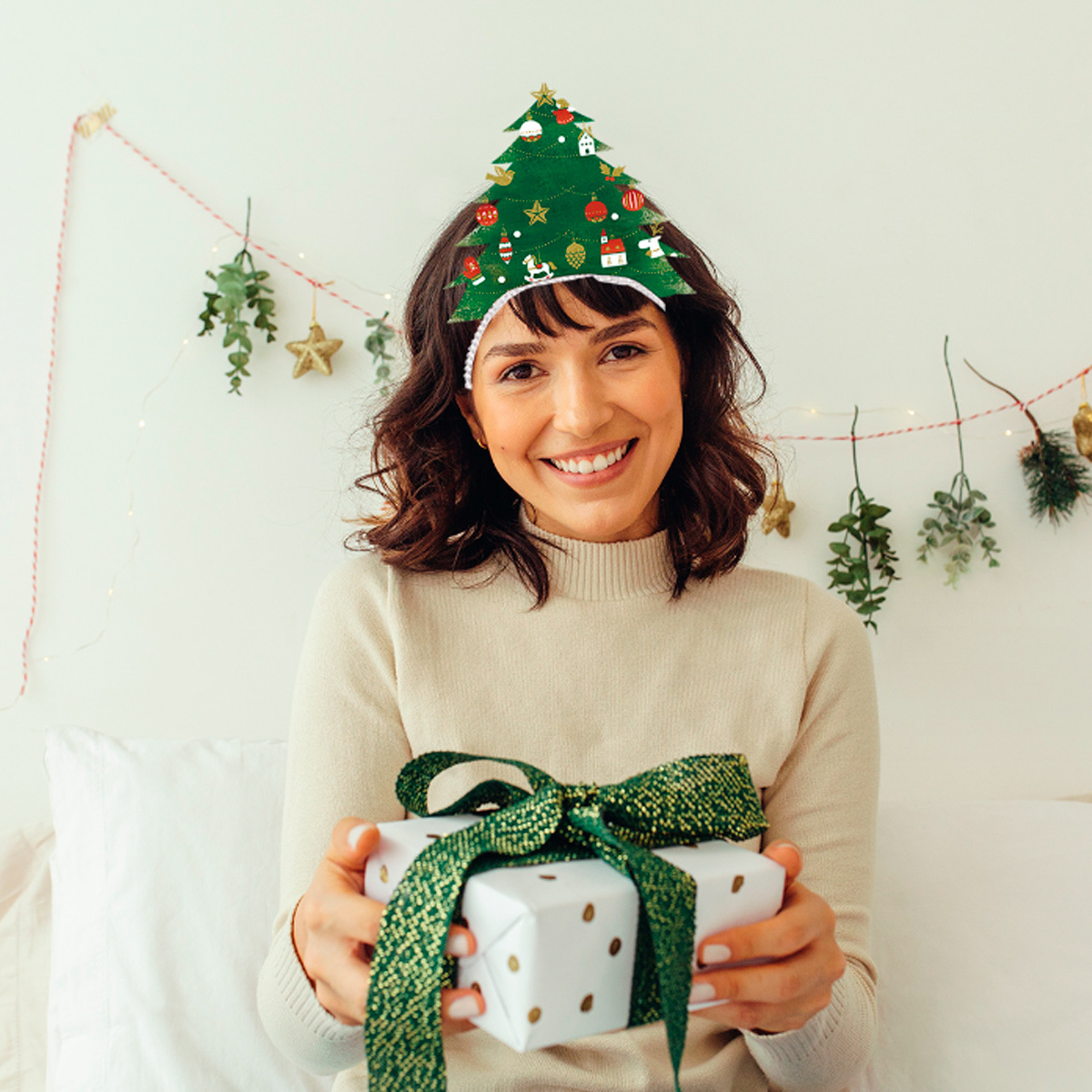 Eine lächelnde Frau hält ein Geschenk. Sie trägt ein Weihnachtsbaum-Stirnband, im Hintergrund ein dekoriertes Zimmer.