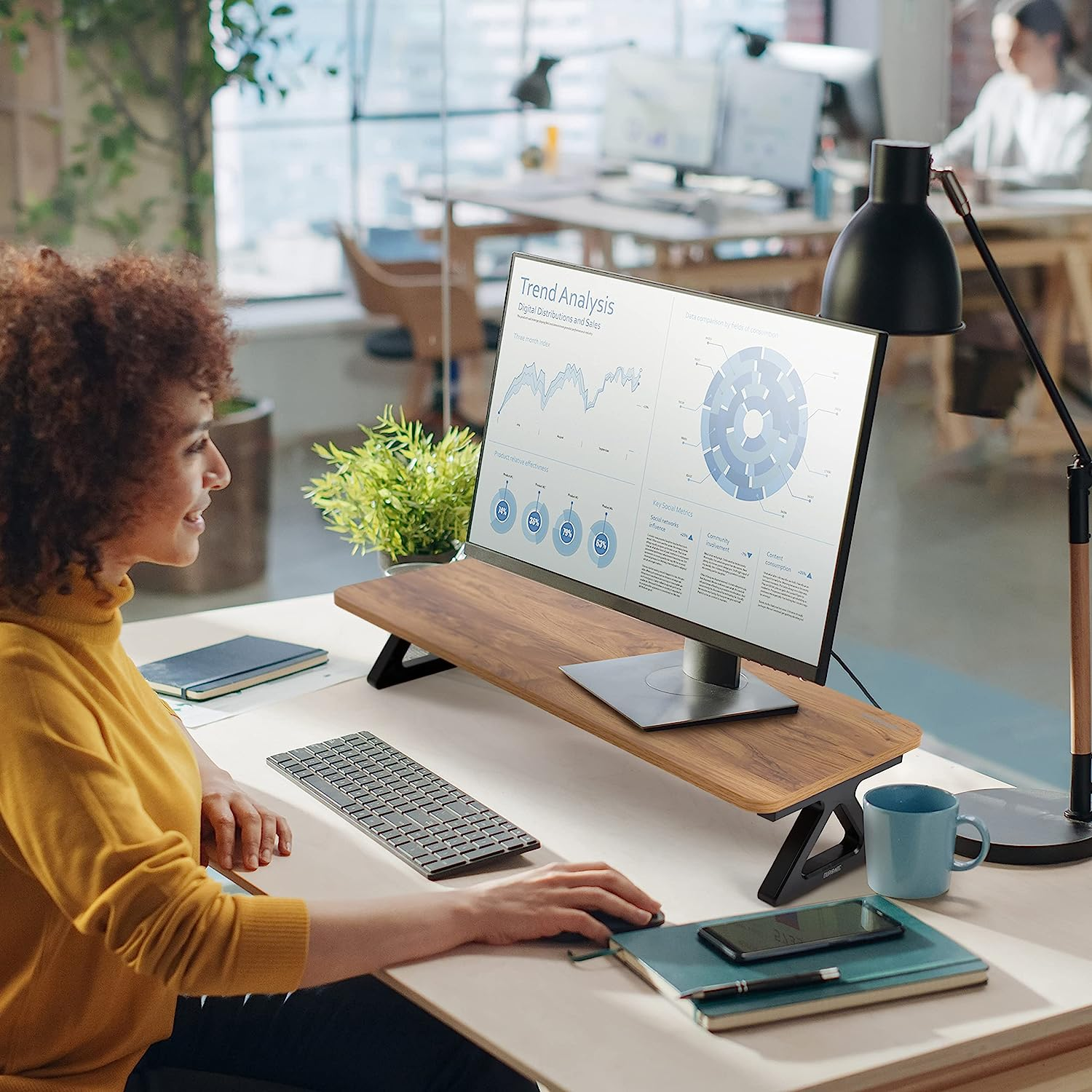 Frau am Schreibtisch mit Monitor auf Ständer. Tastatur, Notizbücher, Becher. Büro Umgebung.