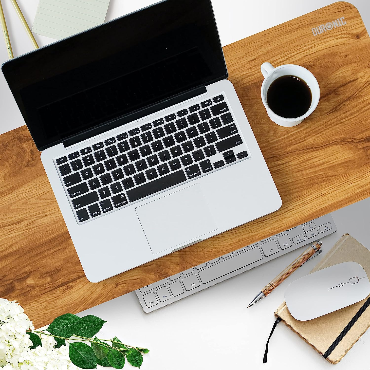 Laptop und Tastatur auf Holzschreibtisch mit Kaffee, Stift, Maus, Notizbuch und Blumen.