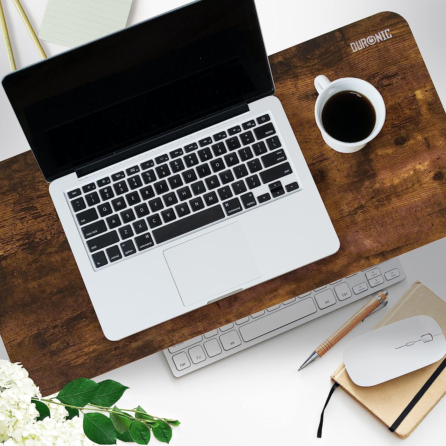 Laptop, Tastatur, Maus und Kaffeetasse auf einem braunen Schreibtisch. Weiße Blumen und ein Notizbuch sind auch zu sehen.