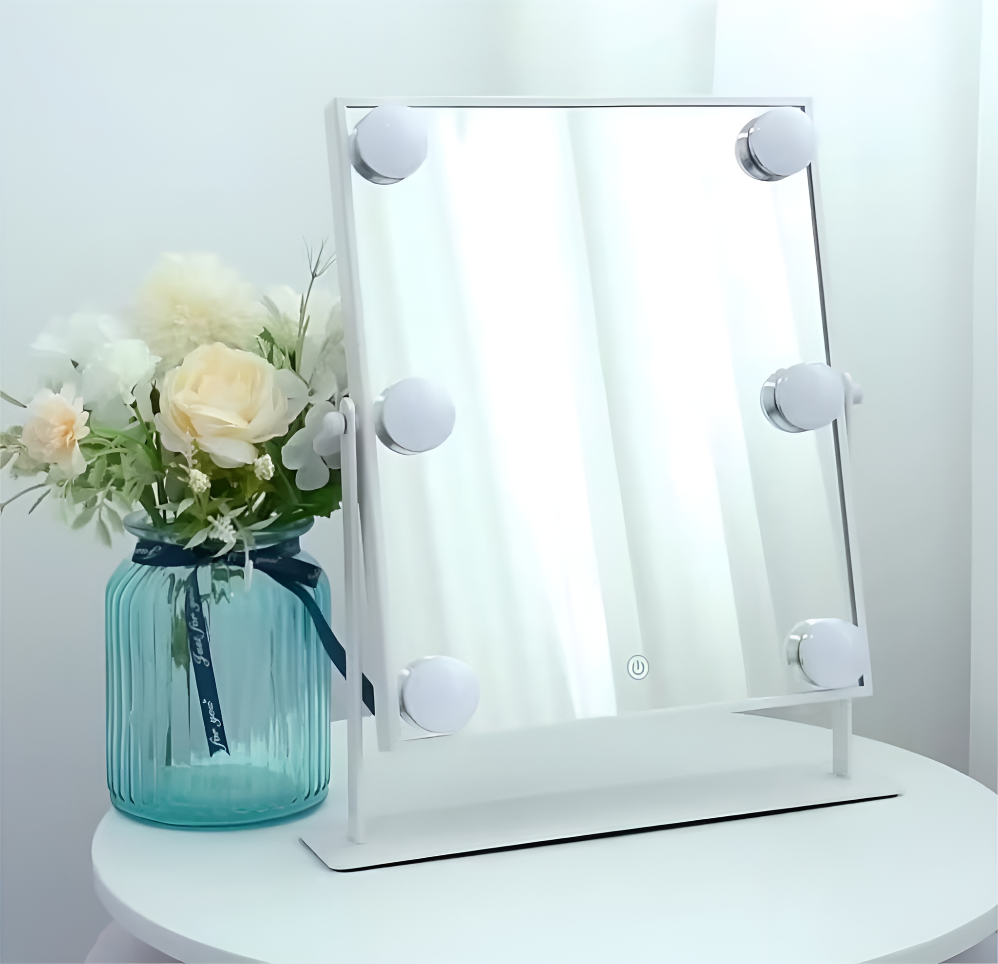 Weißer Schminkspiegel mit sechs Glühbirnen, auf einem Tisch neben einer Vase mit Blumen.