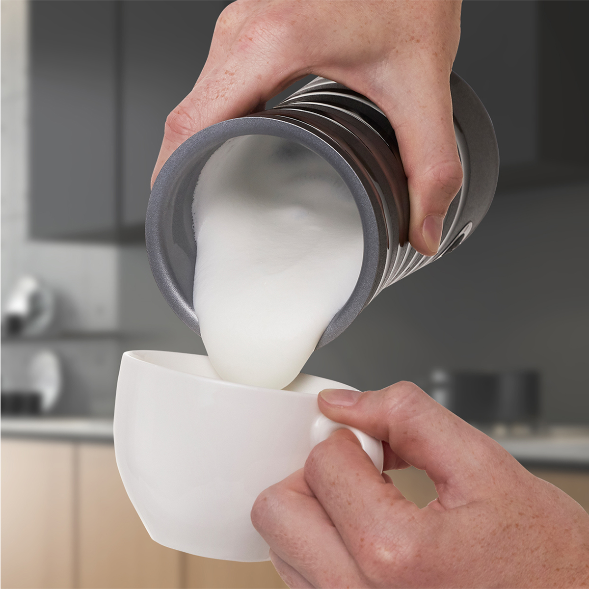 Hände gießen Milch aus einem metallischen Krug in eine weiße Tasse. Hintergrund ist eine Küche.