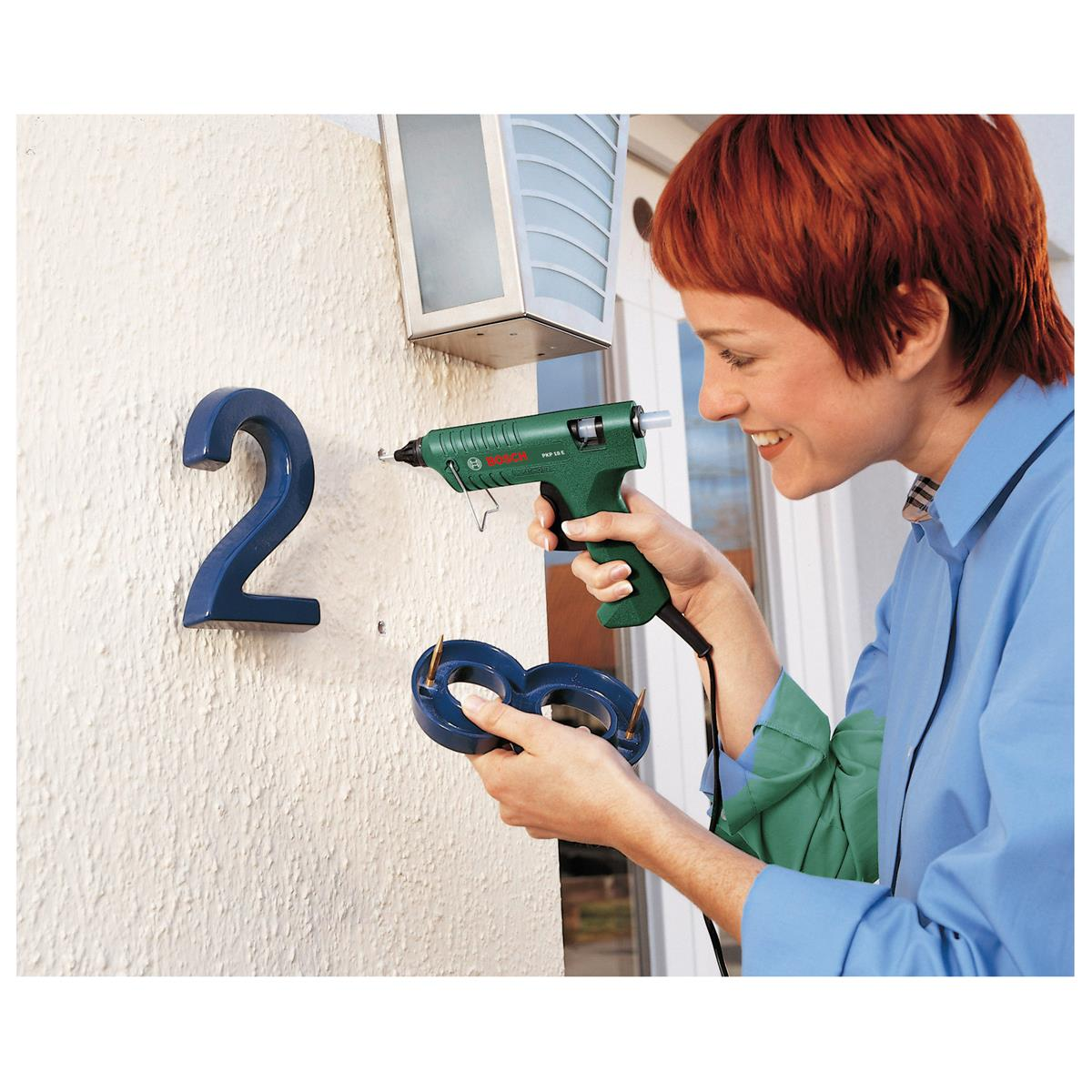 Kobieta przykleja pistoletem do kleju cyfrę 8 do ściany obok niebieskiej cyfry 2.