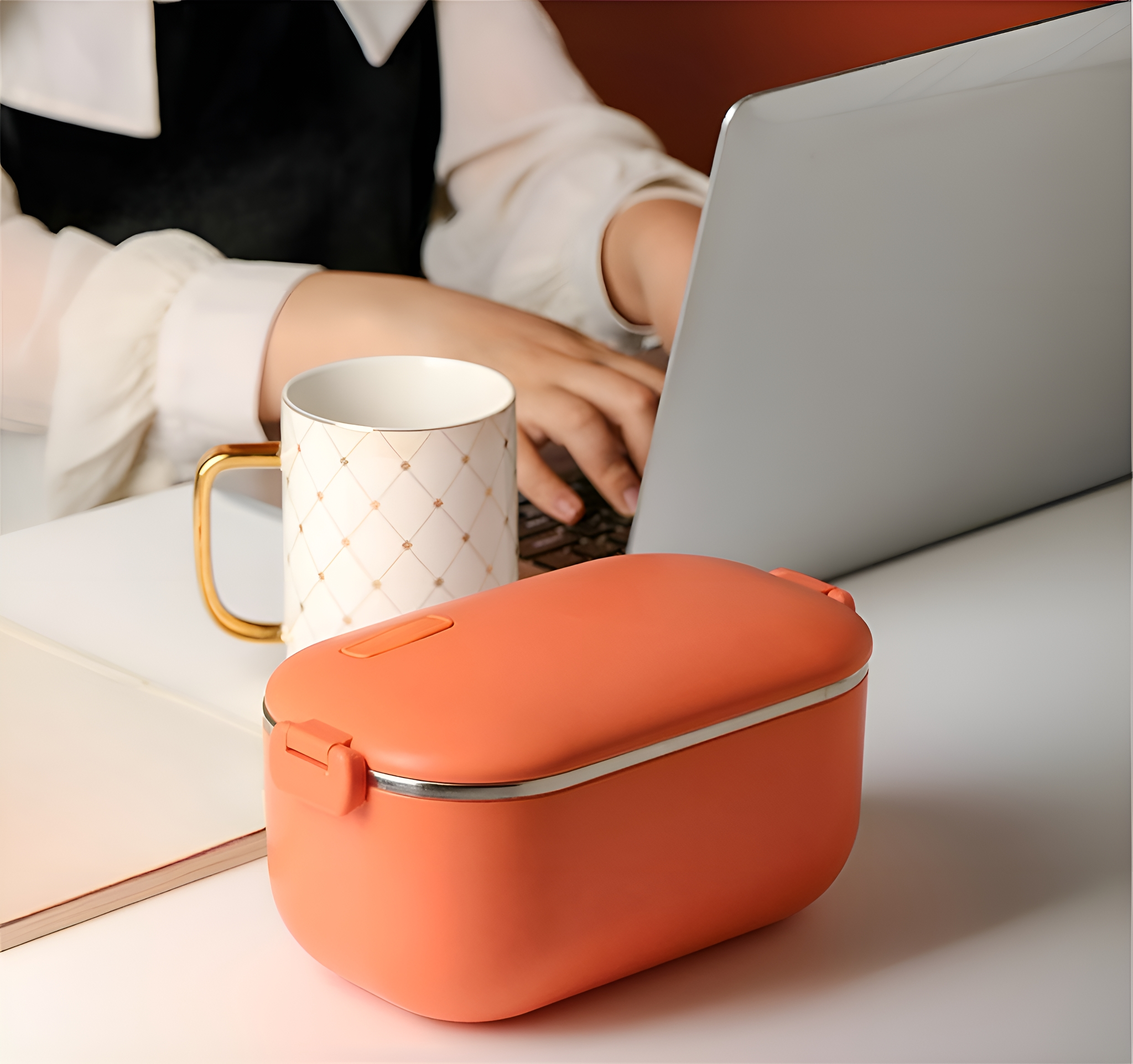 Eine orangefarbene Brotdose mit einem Becher auf einem Tisch. Eine Person tippt im Hintergrund auf einem Laptop.