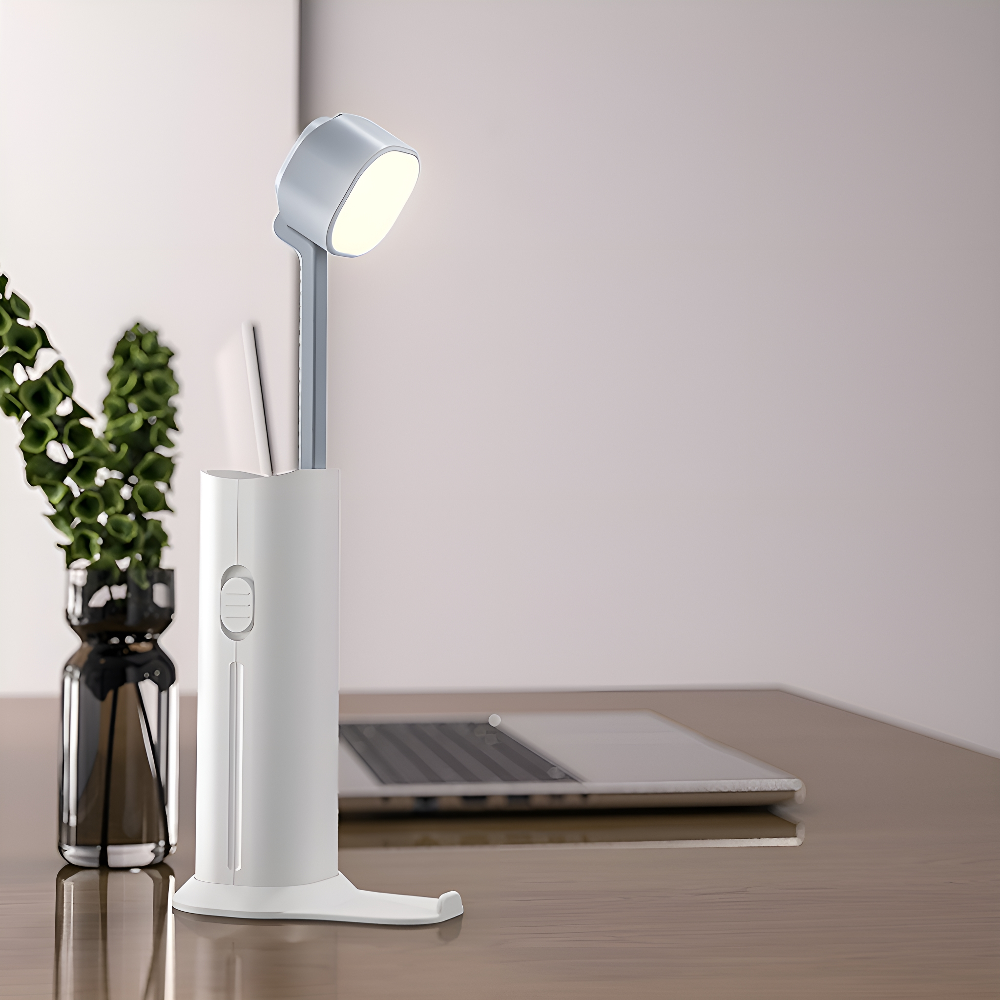 Eine weiße Schreibtischlampe auf einem Holztisch. Ein Laptop und eine Pflanze im Hintergrund.