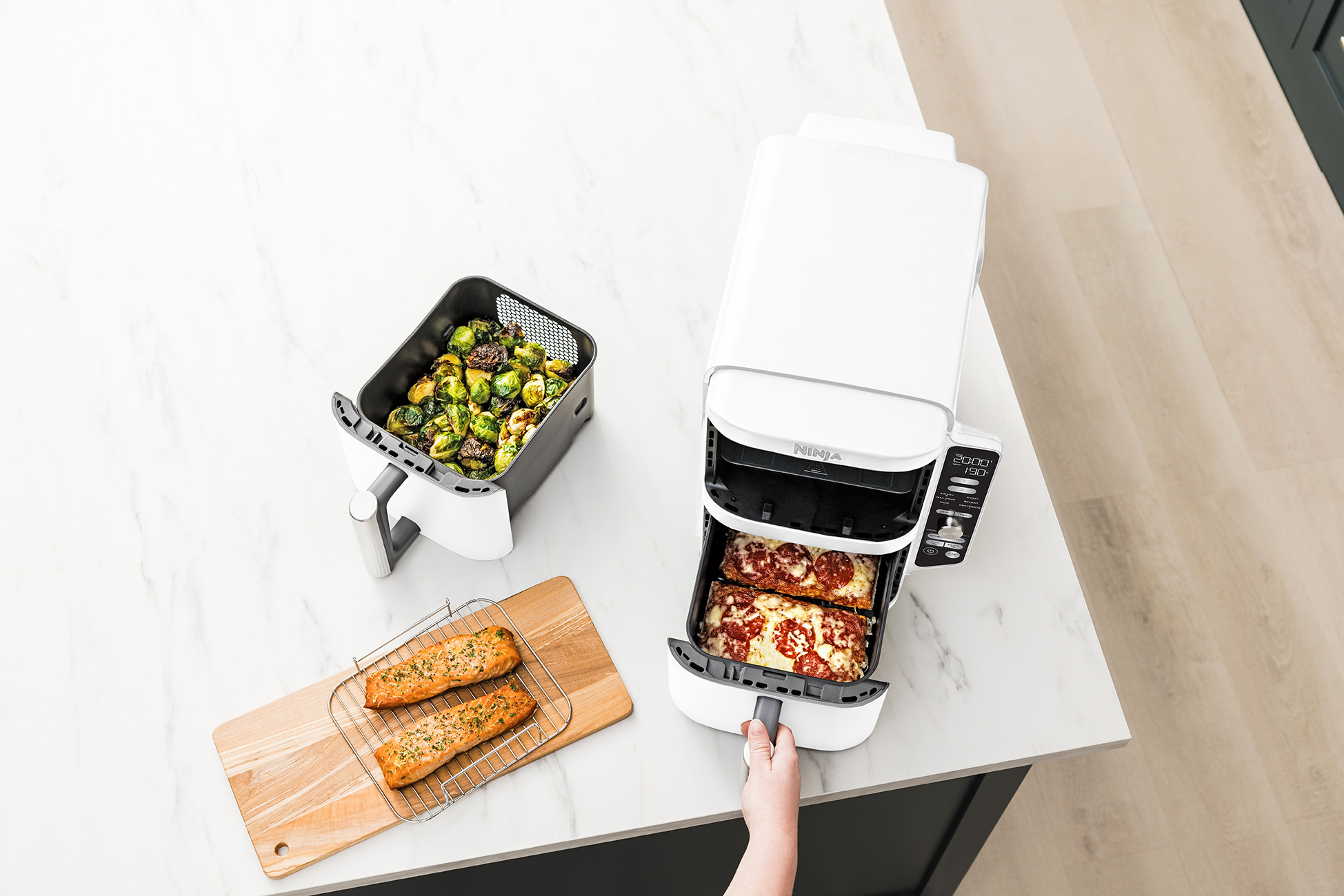 Frytownica beztłuszczowa z jedzeniem. Zawiera upieczoną pizzę, łososia i brukselkę. Osoba wyciąga tacę.