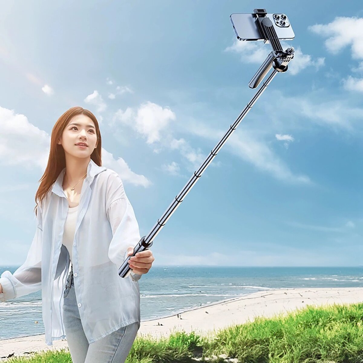 Frau mit Selfie-Stick am Strand. Blauer Himmel und Meer im Hintergrund.