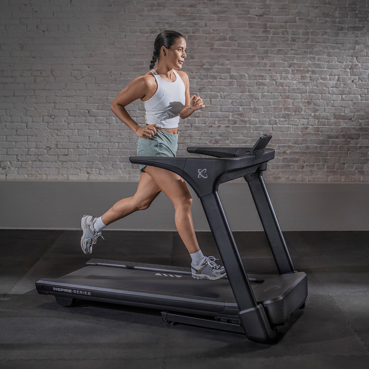 A woman runs on a treadmill. She wears athletic wear and runs in a gym setting with a brick wall.