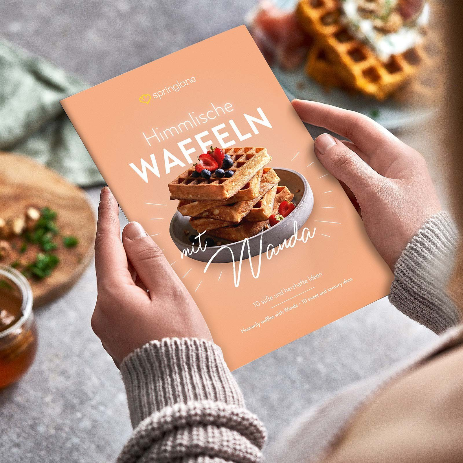 Eine Person hält ein Kochbuch mit dem Titel 'Himmlische Waffeln mit Wanda'. Pfirsichfarbener Einband.