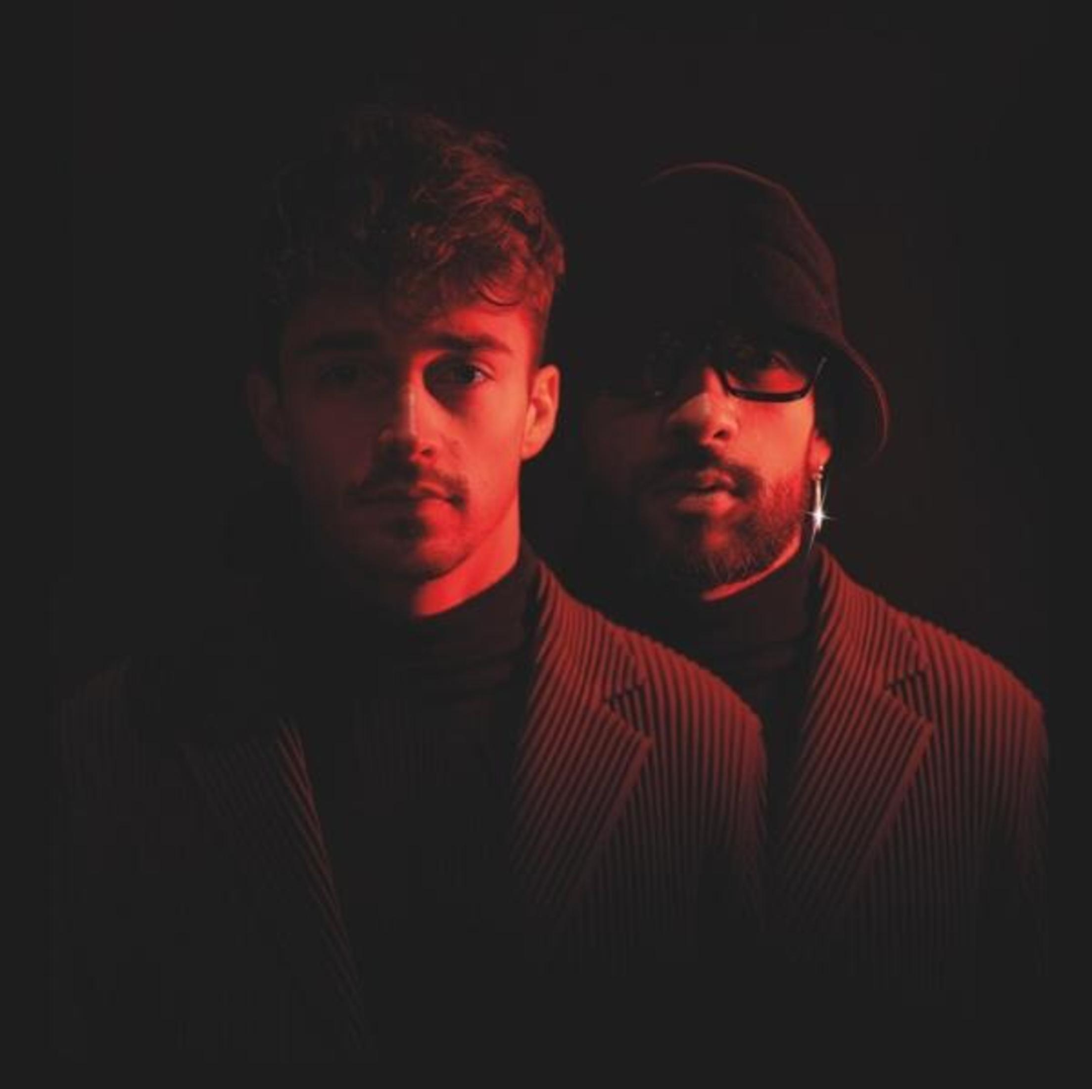 Twee mannen op een donkere achtergrond, naar de kijker gericht. Rood licht verlicht hen. Eén heeft een bril en een hoed.