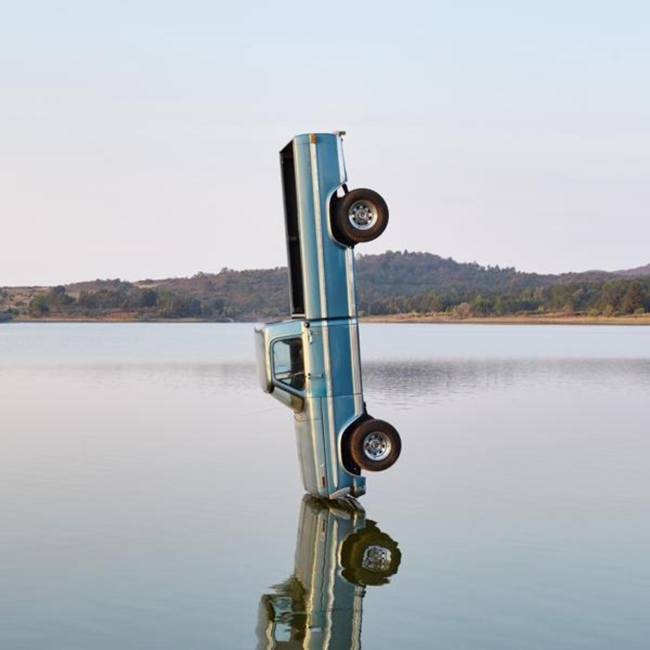Foto: Camioncino azzurro in acqua, capovolto, riflesso. Sfondo cielo.