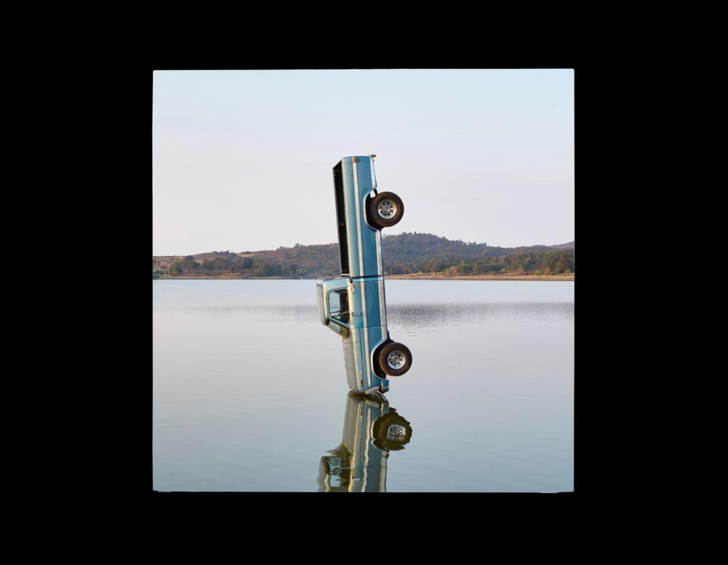 Camioneta azul invertida, reflejada en el agua, con cielo azul y árboles distantes.
