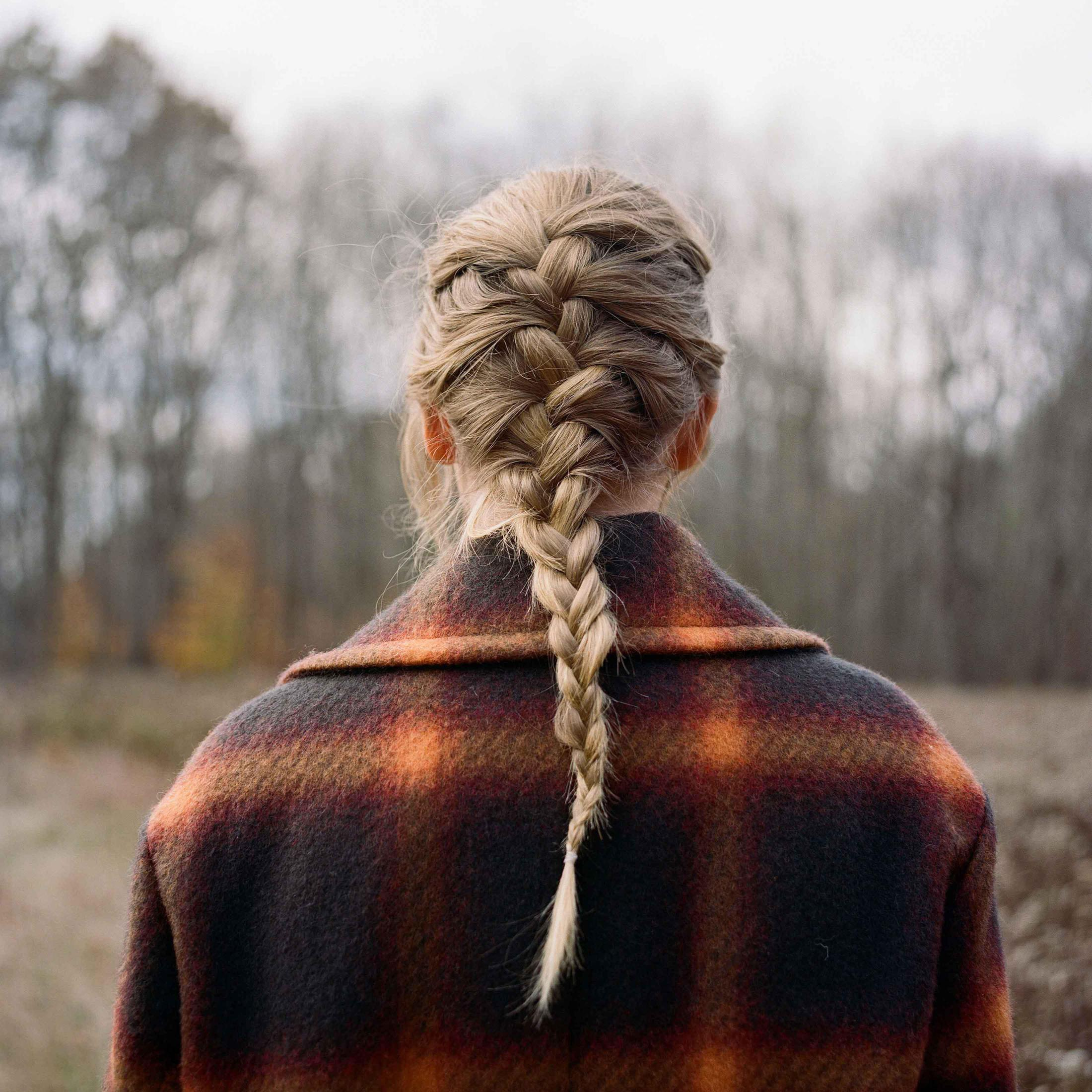 La schiena di una donna con trecce bionde, che indossa un cappotto a scacchi, in un campo con alberi.