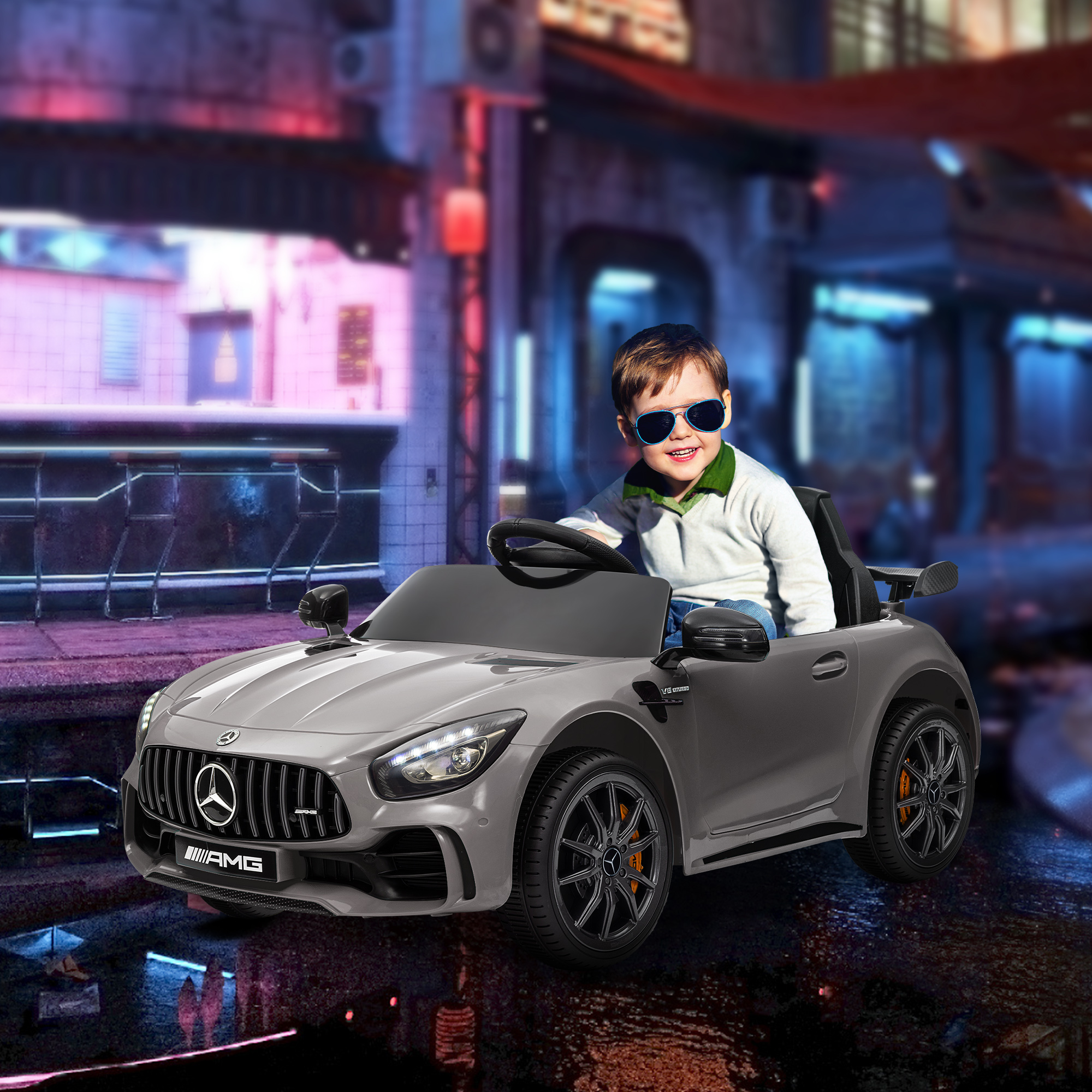 Niño con gafas de sol conduciendo un coche de juguete gris con logo AMG en un entorno urbano.