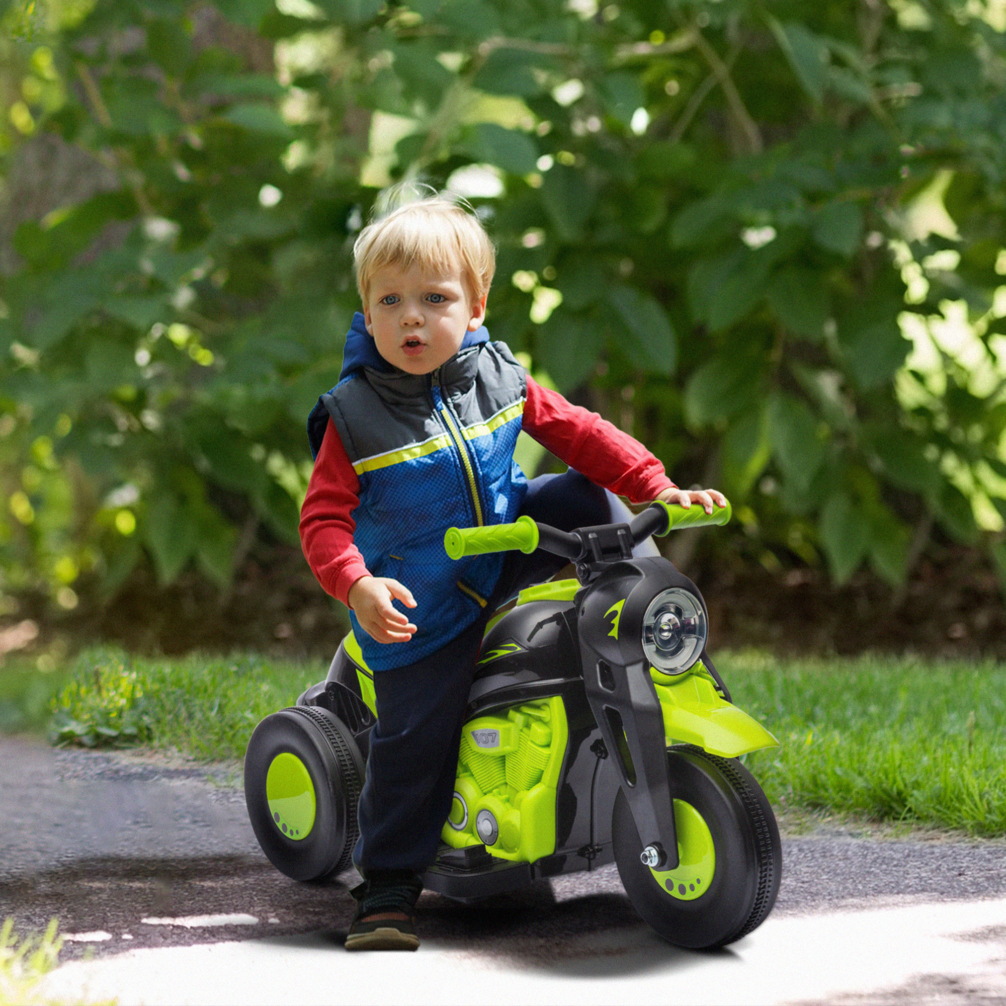 Un niño pequeño está montando en una moto de juguete verde y negra en un camino en un parque. El fondo es verde.