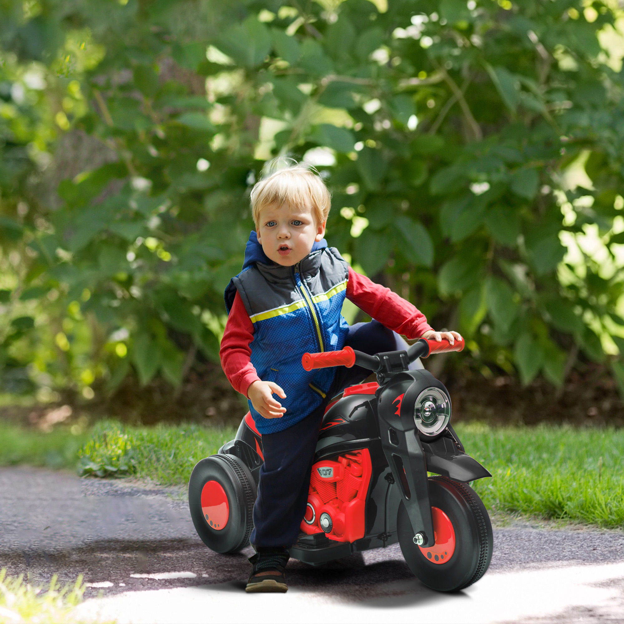 Un niño pequeño en una moto de juguete roja y negra, montando sobre hierba con árboles al fondo.