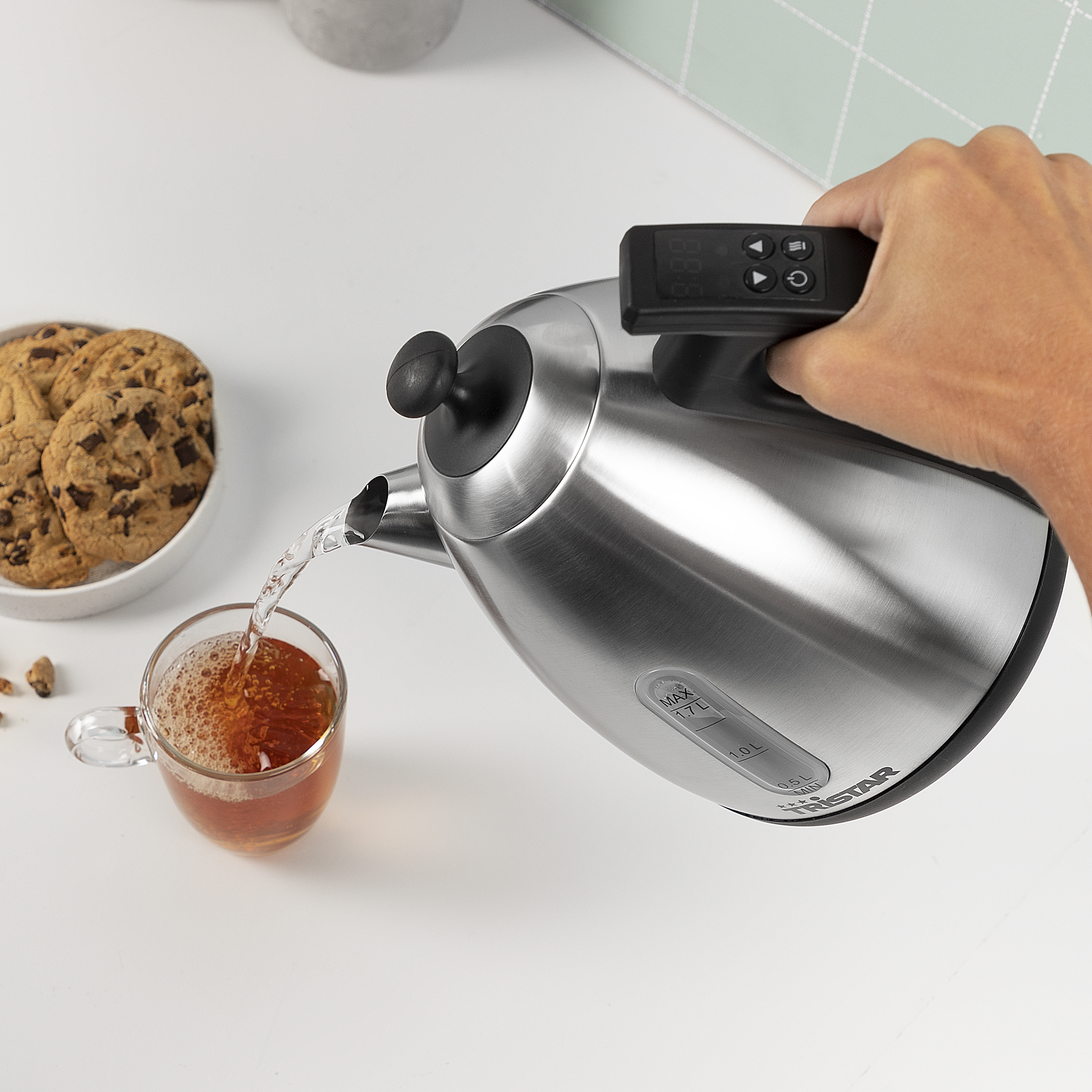 Osoba nalewa wodę ze stalowego czajnika do szklanki herbaty. Ciastka i nakrycie stołu.