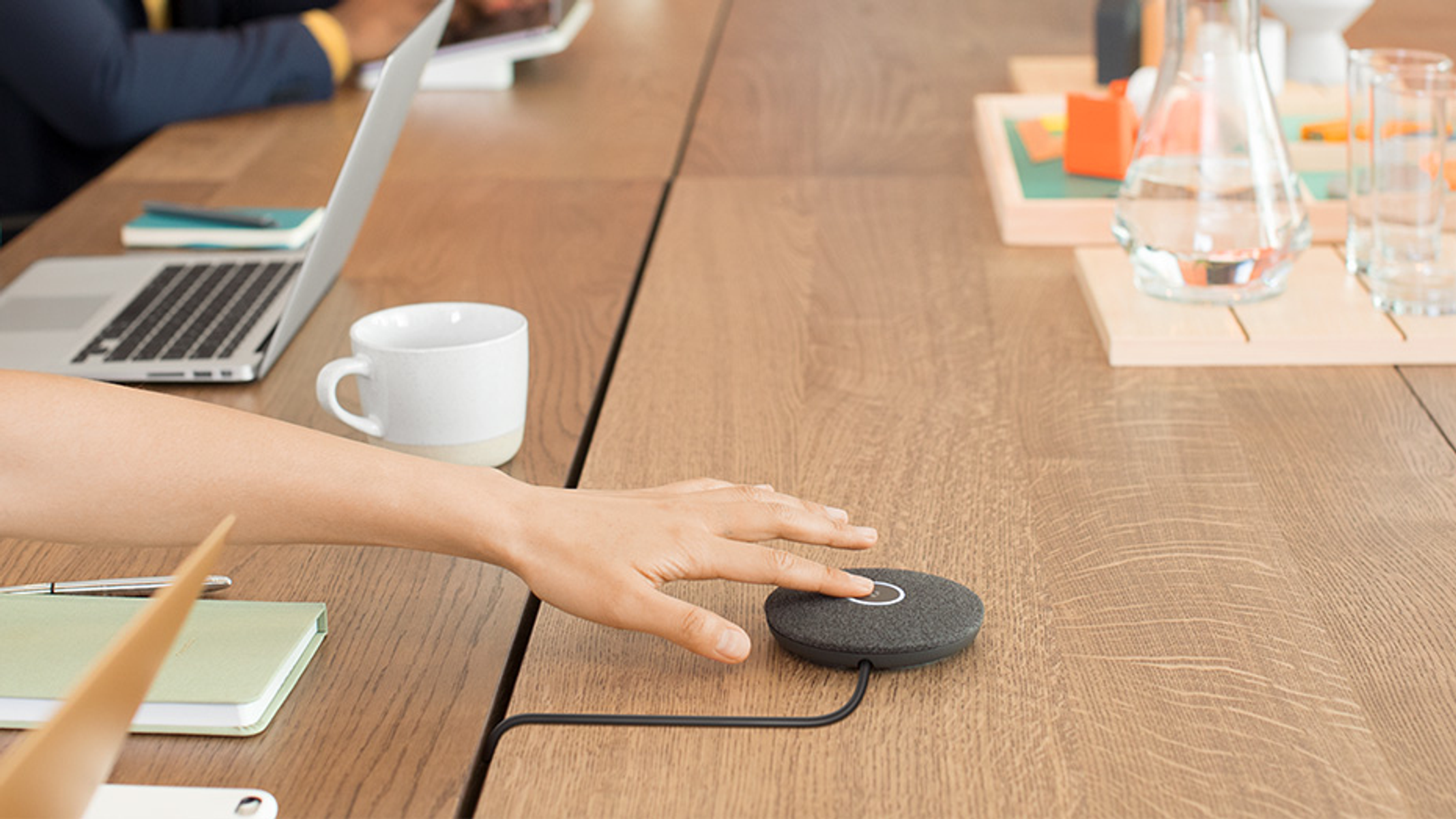 Een hand raakt een zwart rond apparaat aan op een houten tafel. Laptop en mok zijn ook aanwezig.