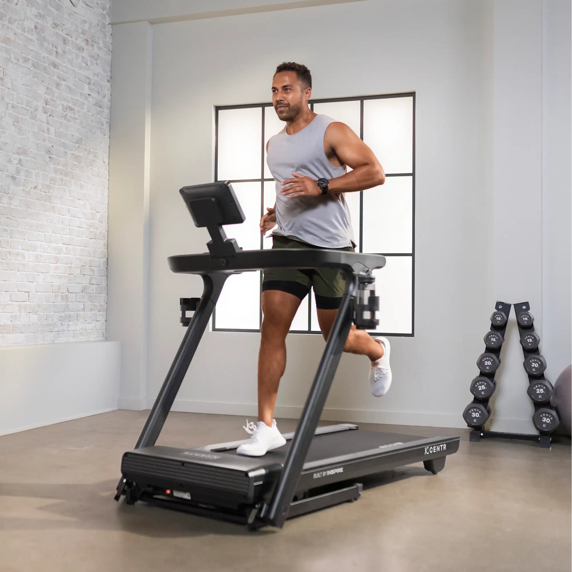 A man running on a black treadmill in a well-lit room with weights in the background.