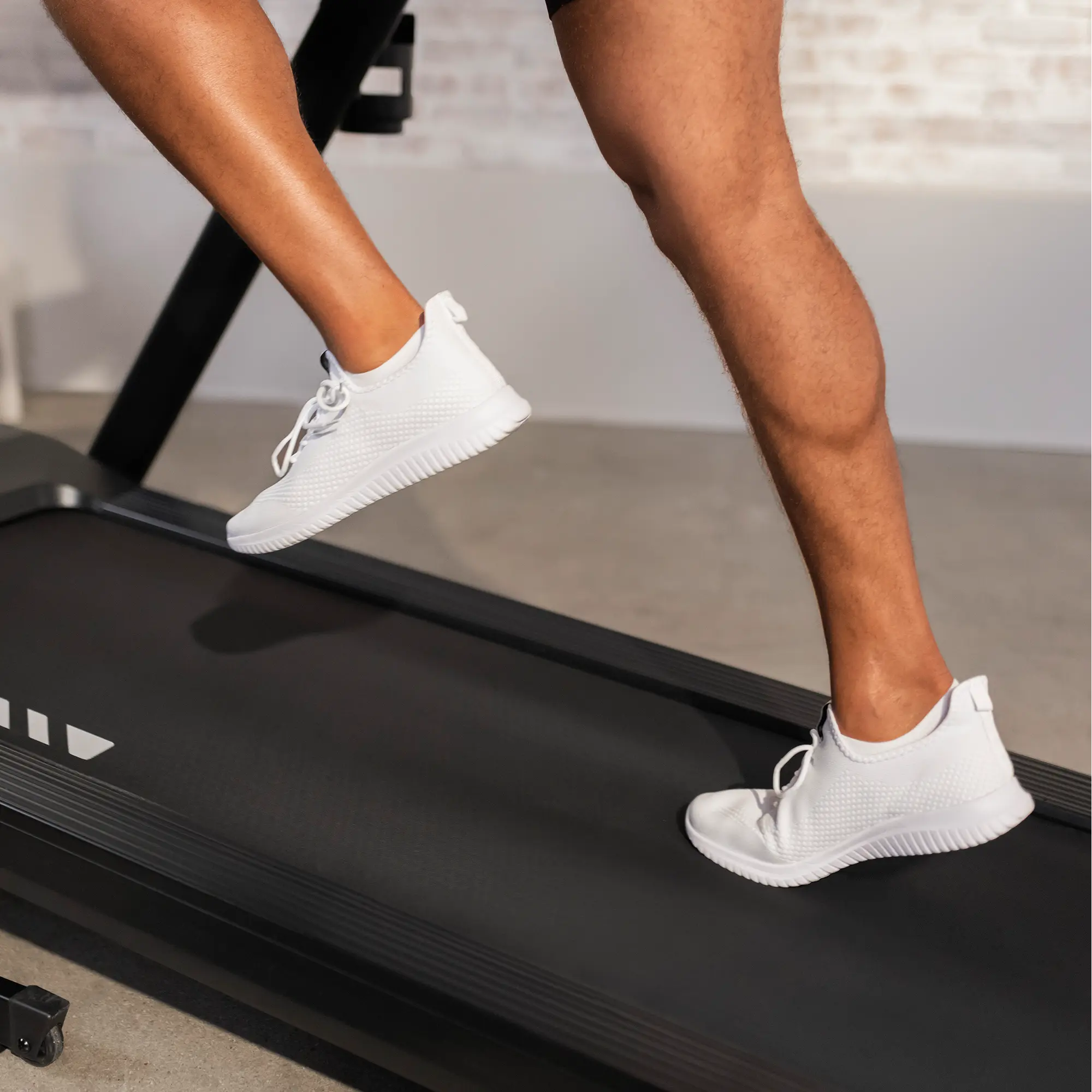 A person's legs, wearing white sneakers, running on a black treadmill. The background is a wall.
