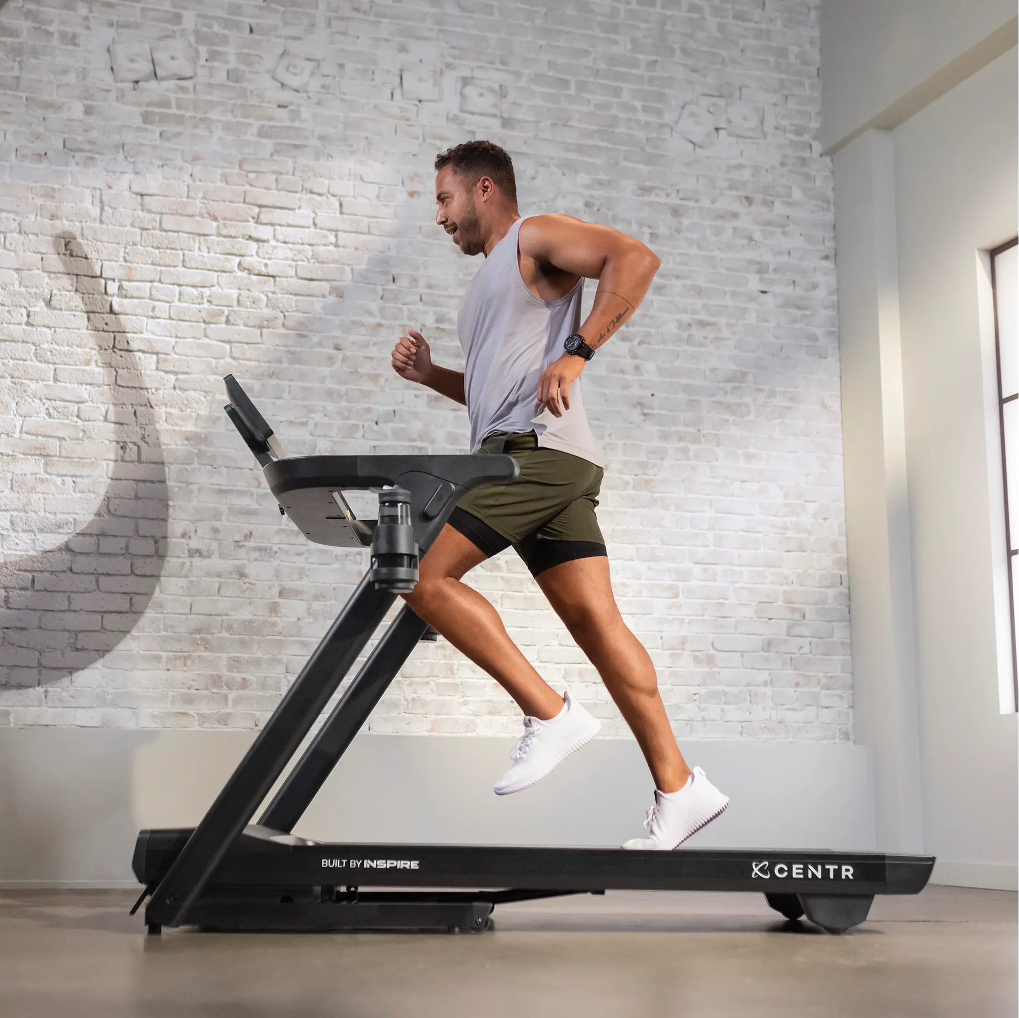 Man running on a black treadmill in a gym with a brick wall background.