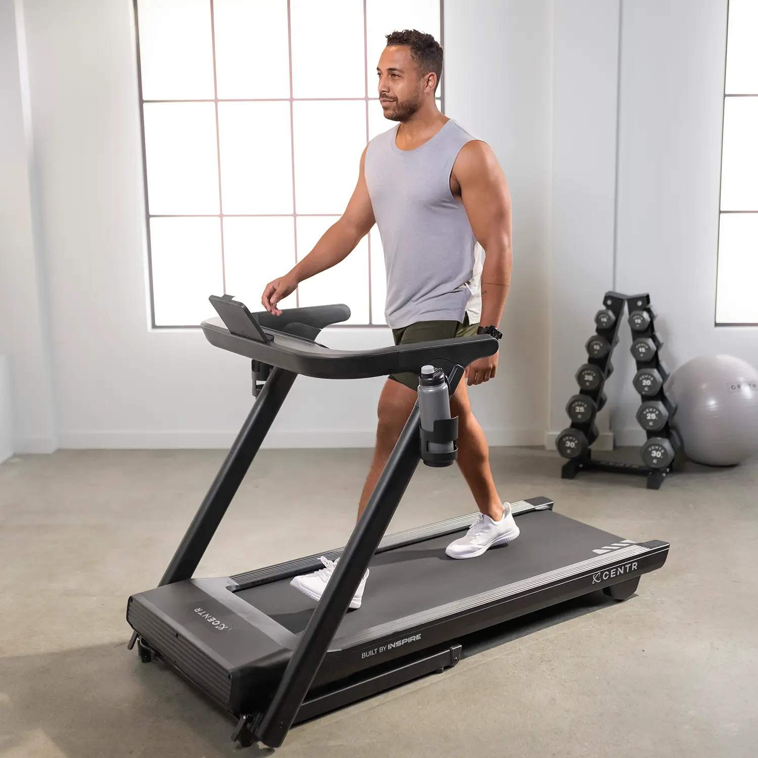Man on a treadmill, wearing a gray tank top and shorts. There is a dumbbell rack and ball.