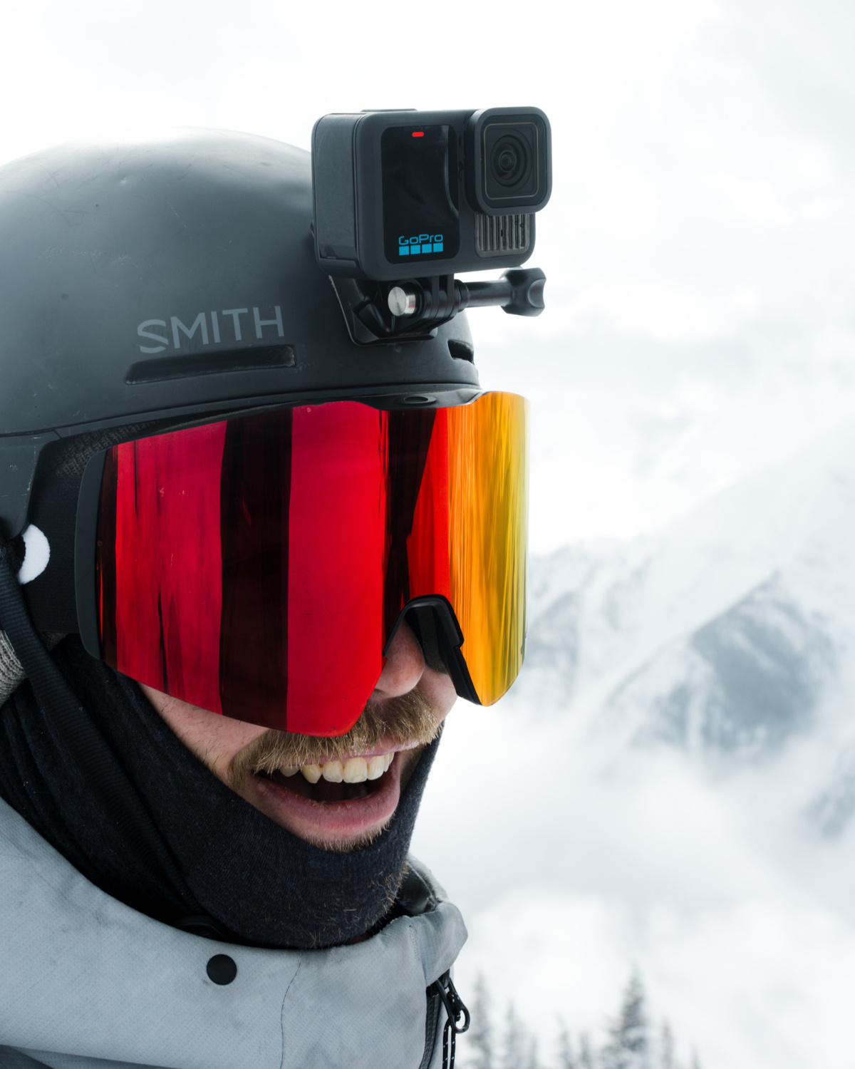 Skier with goggles and a GoPro on his helmet, smiling. Snowy mountains in the background.
