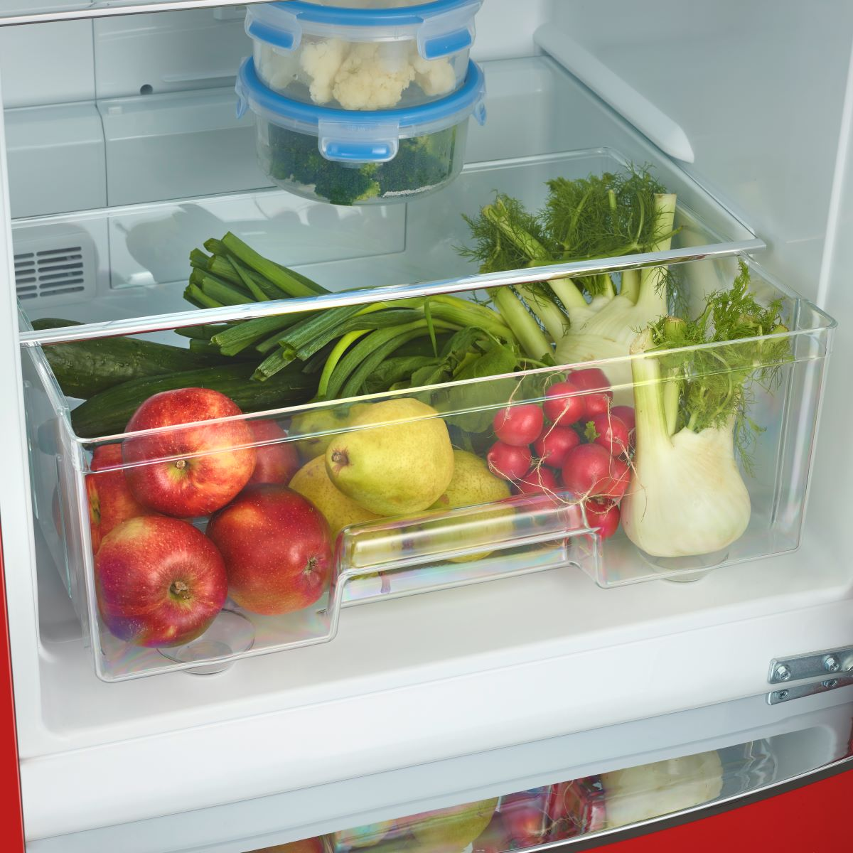 Wnętrze lodówki z różnymi warzywami i owocami w przezroczystych pojemnikach i szufladach.