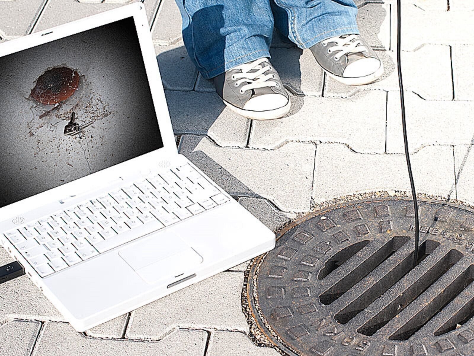 Ein Laptop neben einem offenen Kanaldeckel, Beine und Schuhe einer Person sichtbar.