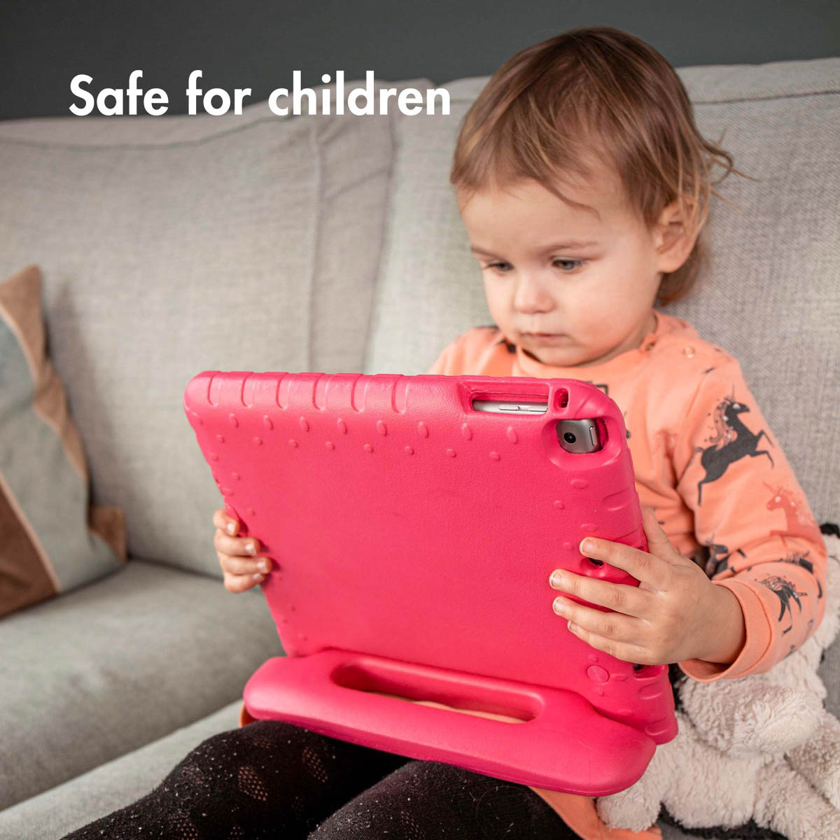 Un tout-petit tenant une tablette rose dans un étui rose. L'enfant est sur un canapé et porte une chemise orange.