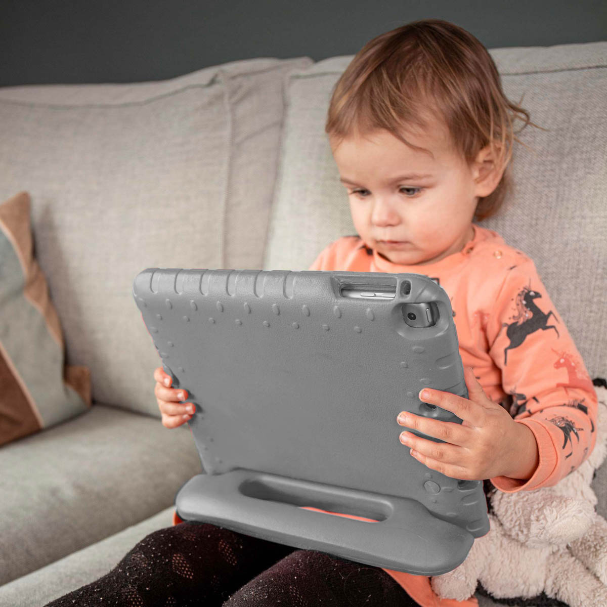 Een peuter zit en houdt een tablet vast in een grijze hoes. Het kind kijkt naar het scherm.