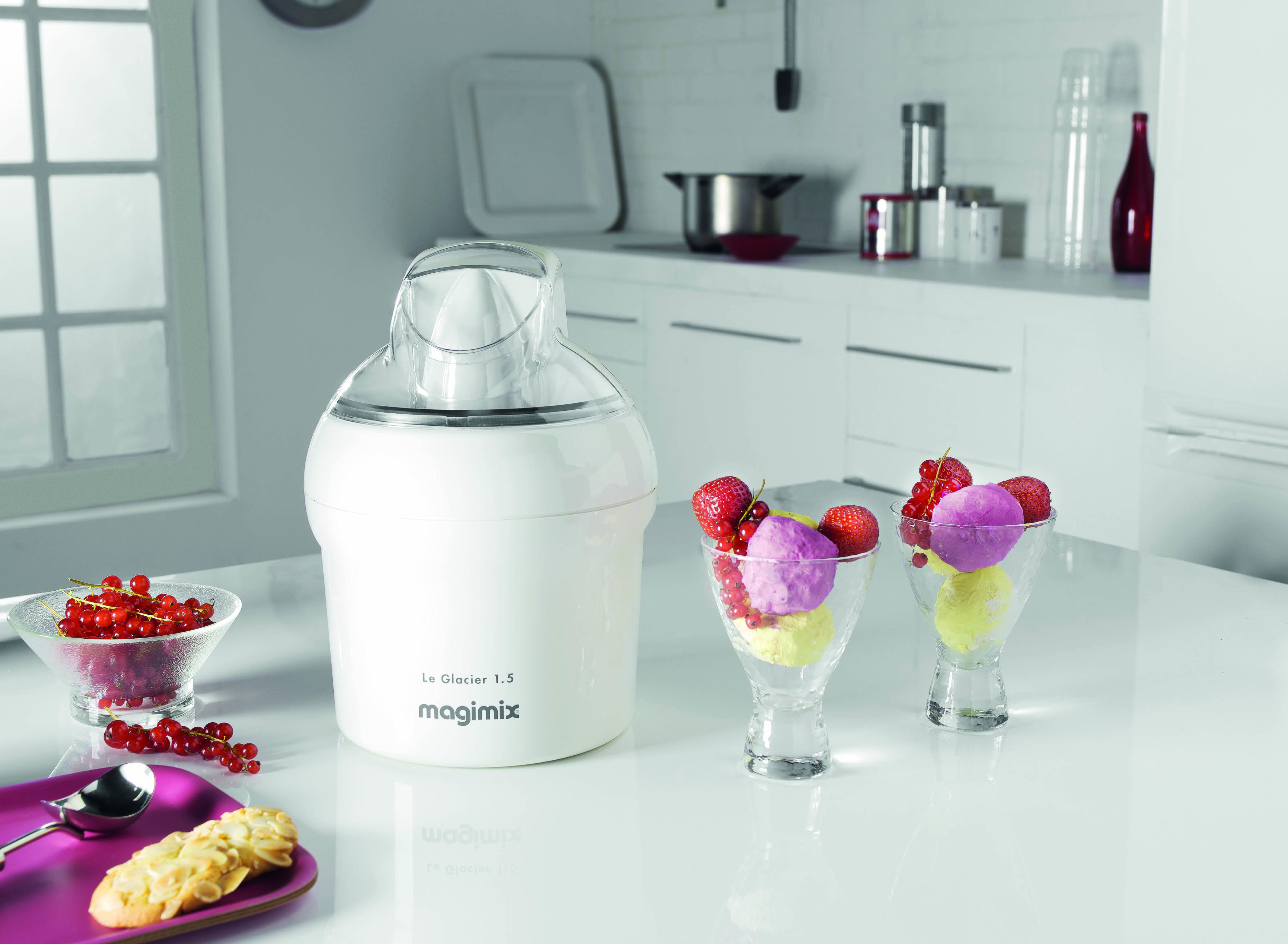 A white ice cream maker with ice cream in glasses on a table. Red currants and cookies are also on the table.