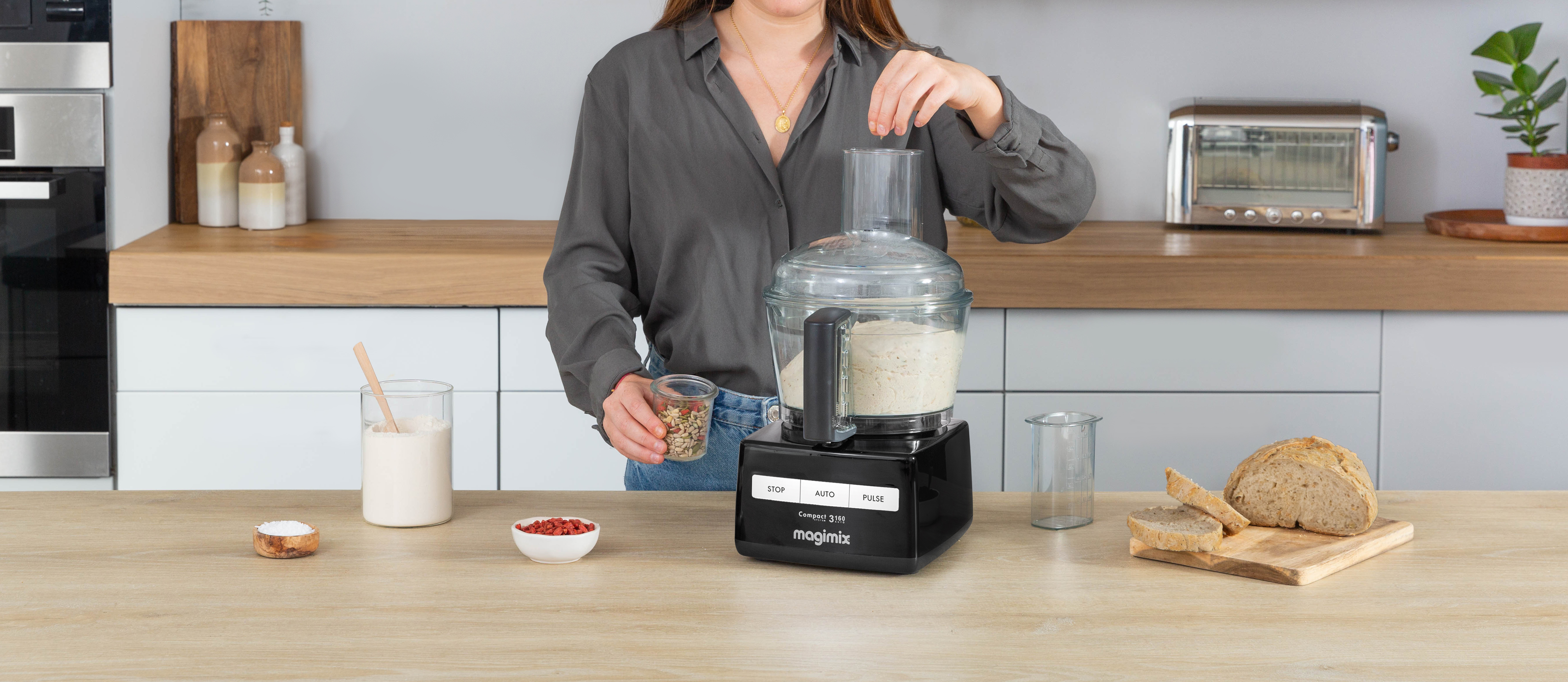 Vrouw voegt ingrediënten toe aan een keukenmachine. Diverse etenswaren op een houten aanrecht.